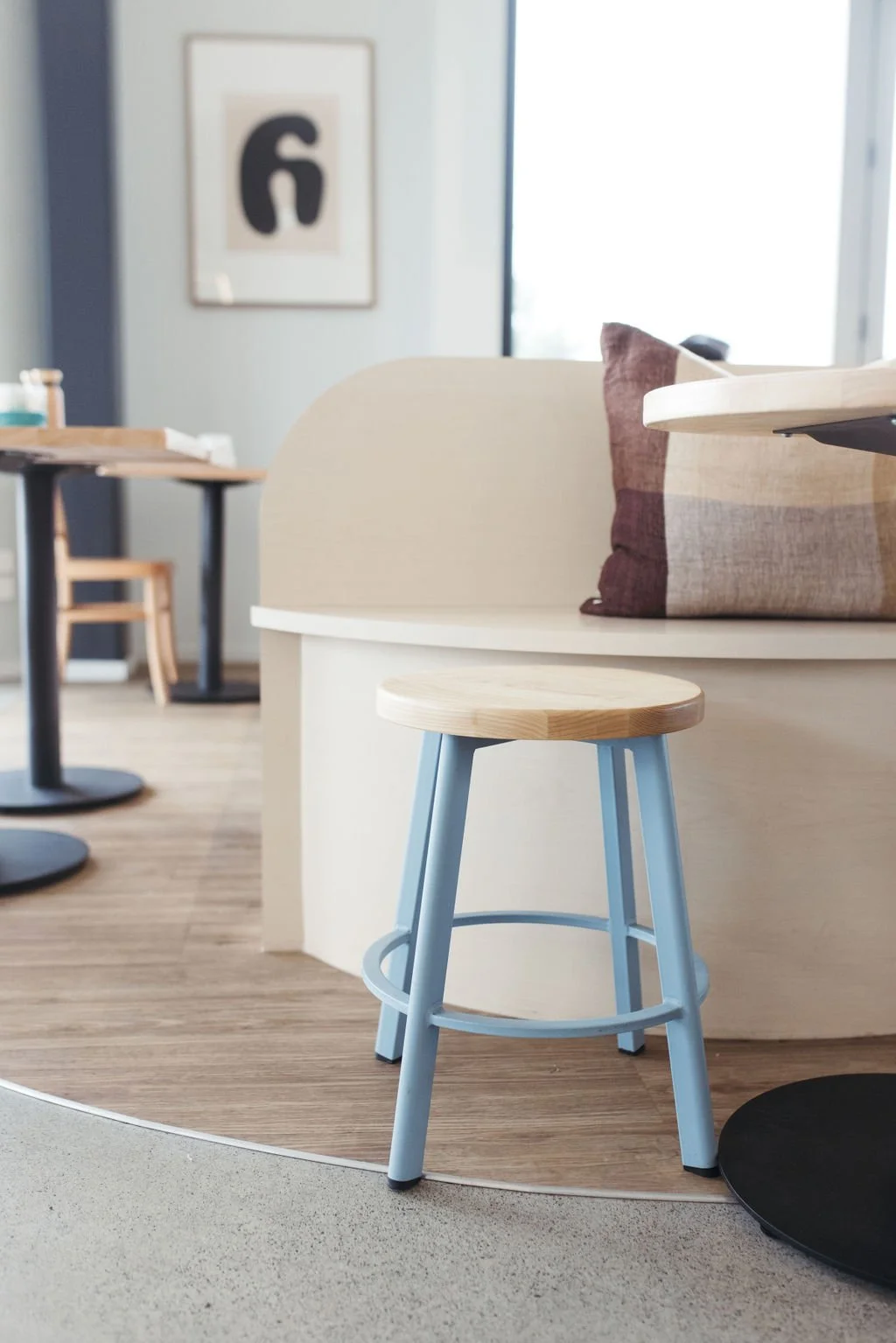 Indoor space featuring a small wooden stool with light blue legs in the foreground. In the background, there are tables, chairs, a sofa with a purple and beige pillow, and abstract wall art. Natural light is coming through a large window.