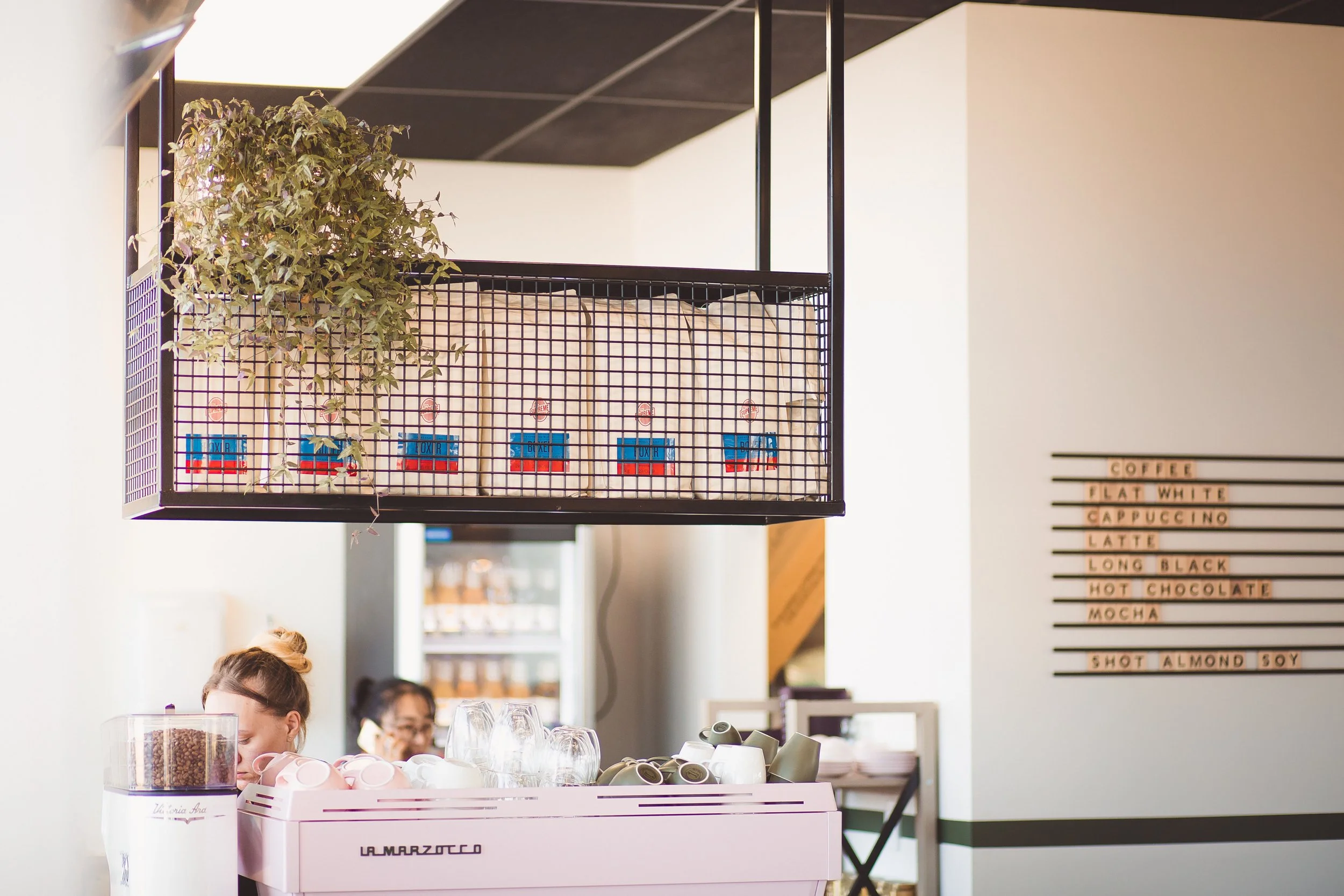 Inside a coffee shop with a barista preparing drinks behind a pink La Marzocco espresso machine. There is a menu on the wall listing coffee options like flat white, cappuccino, latte, long black, hot chocolate, mocha, and a shot almond soy. A hanging shelf with packages and a plant is above the counter.