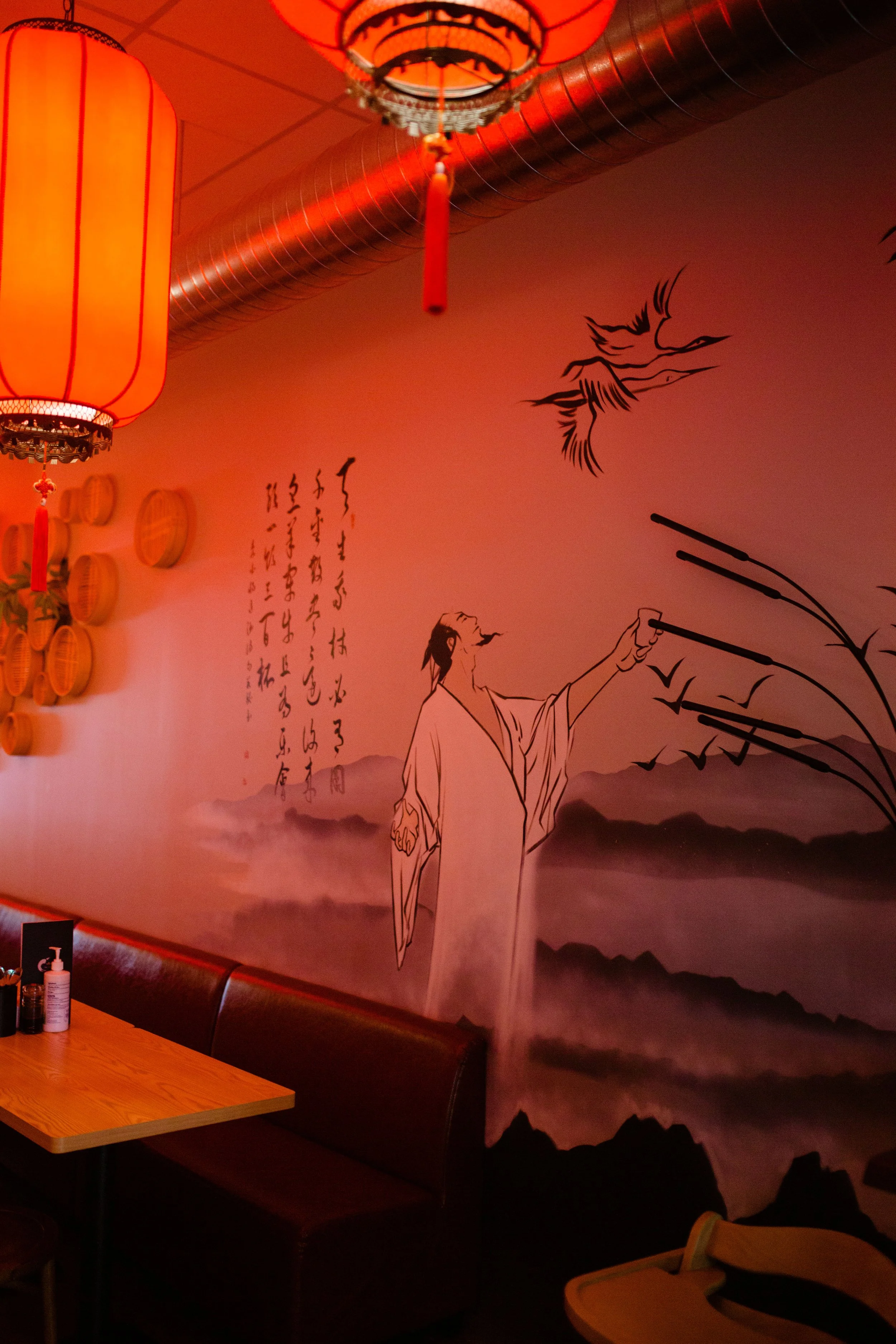 Decorative wall mural featuring a traditional Chinese painting style with a man in flowing robes reaching out to flying birds, mountains in the background, Chinese calligraphy, and hanging lanterns in a restaurant.