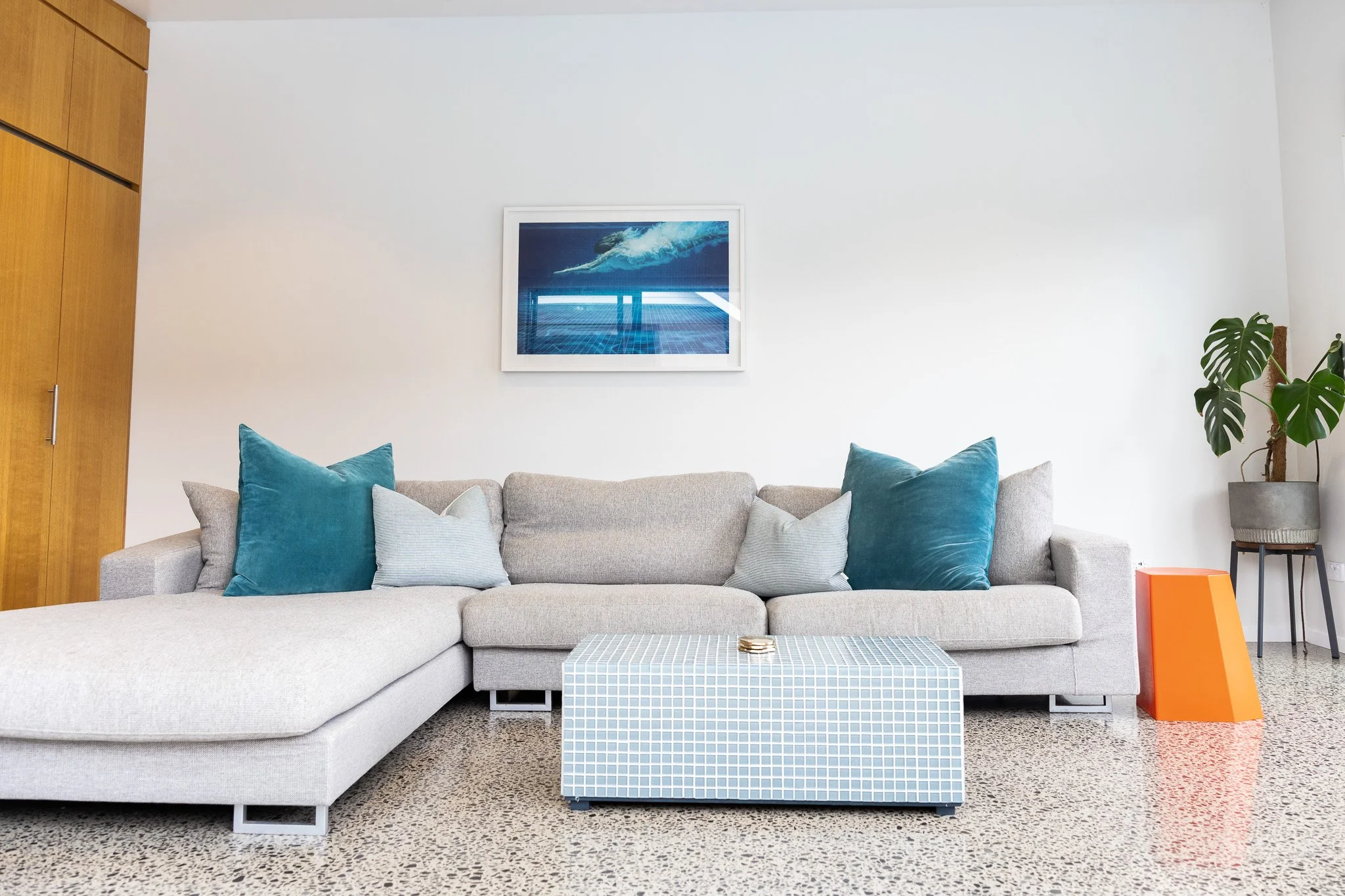 Living room with a light gray sectional sofa accented with blue and white pillows, a patterned ottoman, a potted plant in a gray planter, an orange side table, a framed aquatic photograph, and a wooden built-in cabinet on the left side of the image.