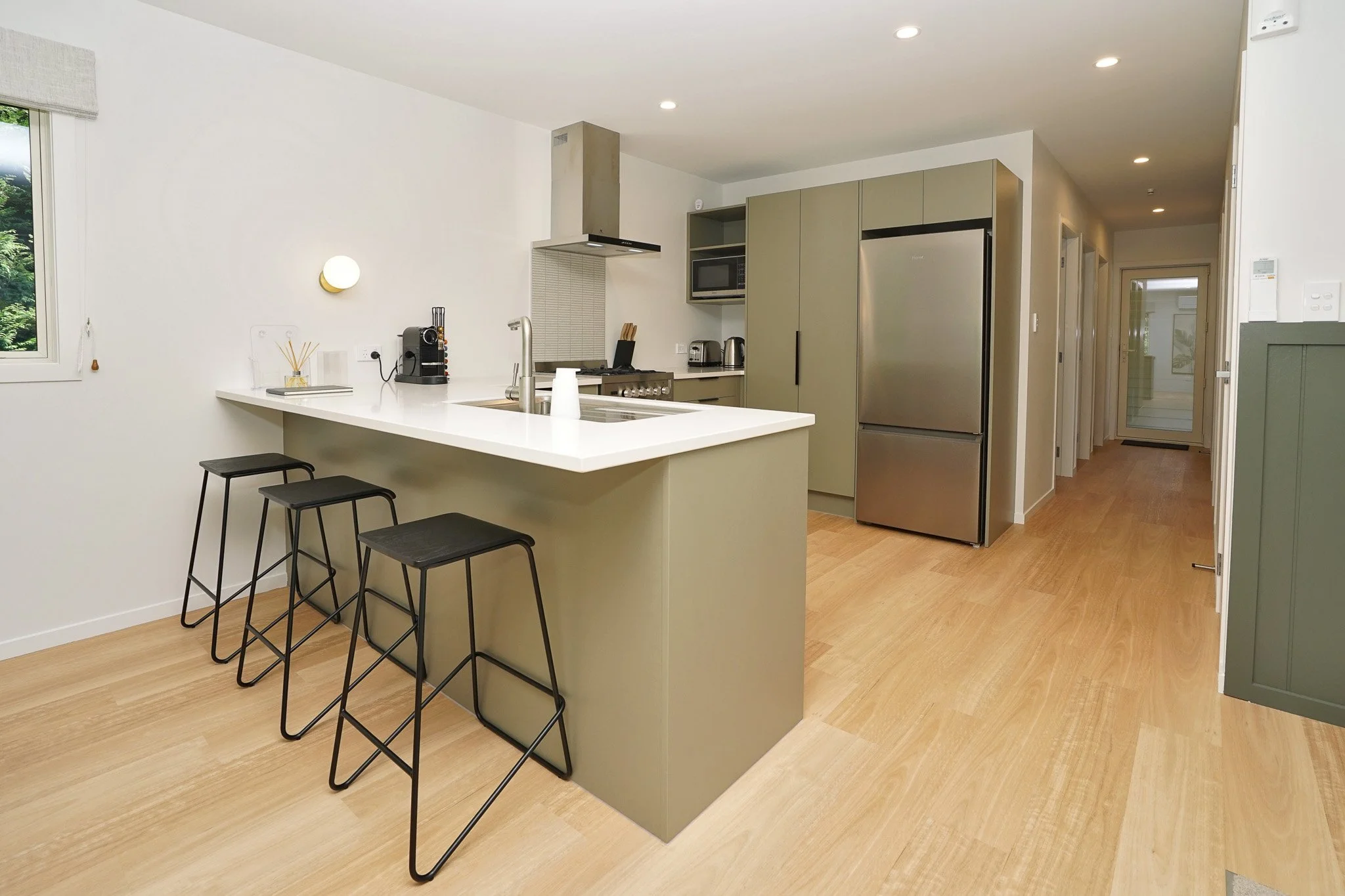 Modern kitchen with light wood flooring, beige walls, gray cabinets, stainless steel refrigerator, white countertop with bar stools, small window, and hallway leading to other rooms.