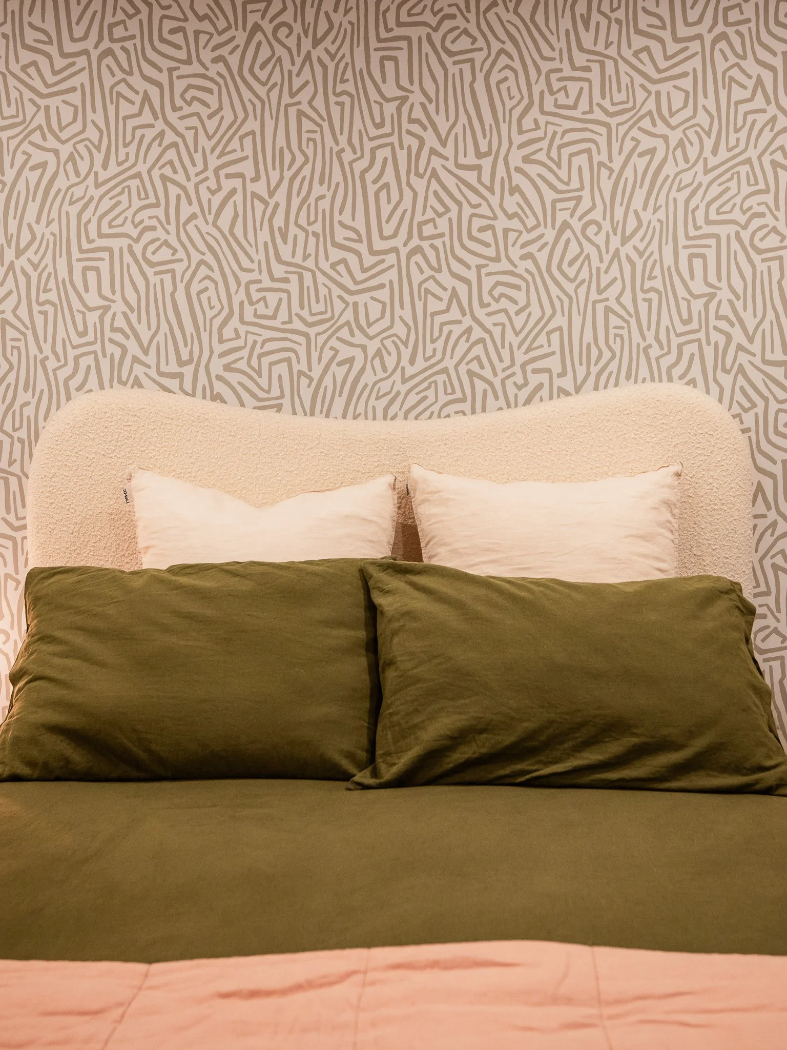 A neatly made bed with an olive-green bedspread, two matching pillows, and two white pillows against a textured off-white headboard, with a patterned wallpaper in shades of beige and taupe in the background.