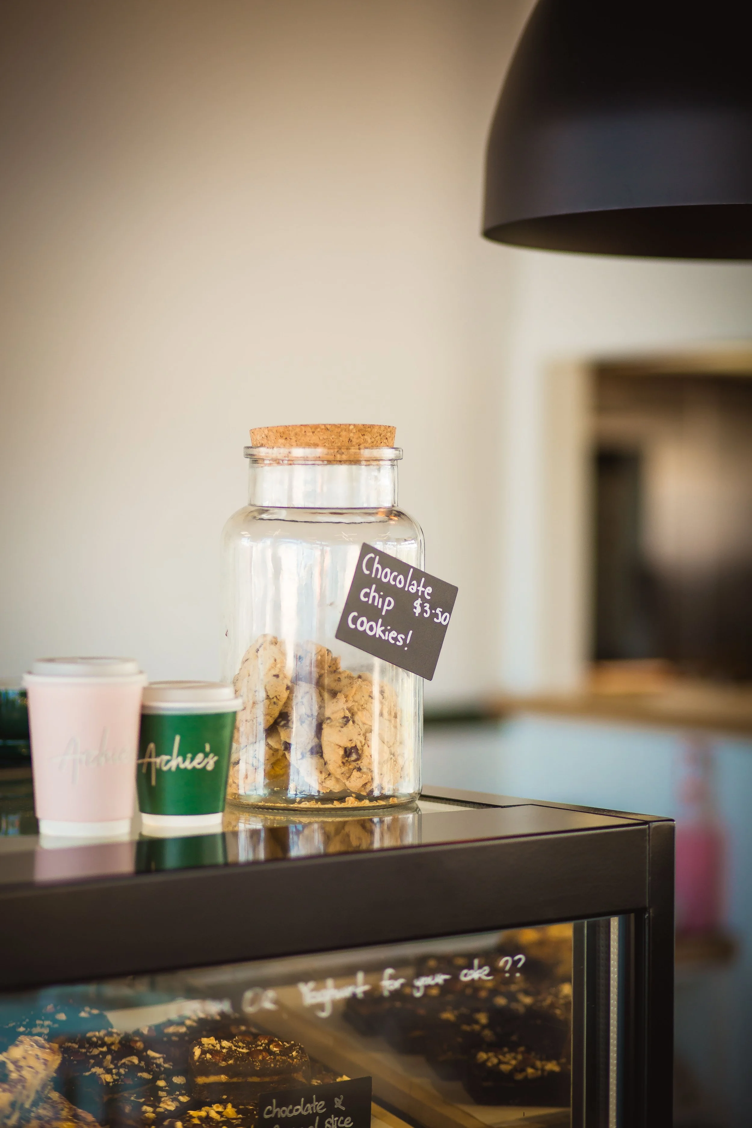 A glass jar with a cork lid containing chocolate chip cookies on a counter at a café, with a small chalkboard sign indicating the cookies cost $3.50.