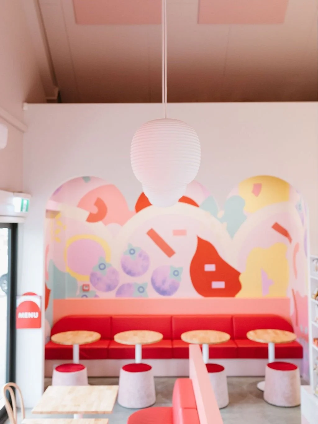Indoor seating area with red cushioned bench, four small round tables with red and white bases, pastel abstract wall mural, hanging white paper lantern light, door with an emergency exit sign, and partially visible menu sign on the left.