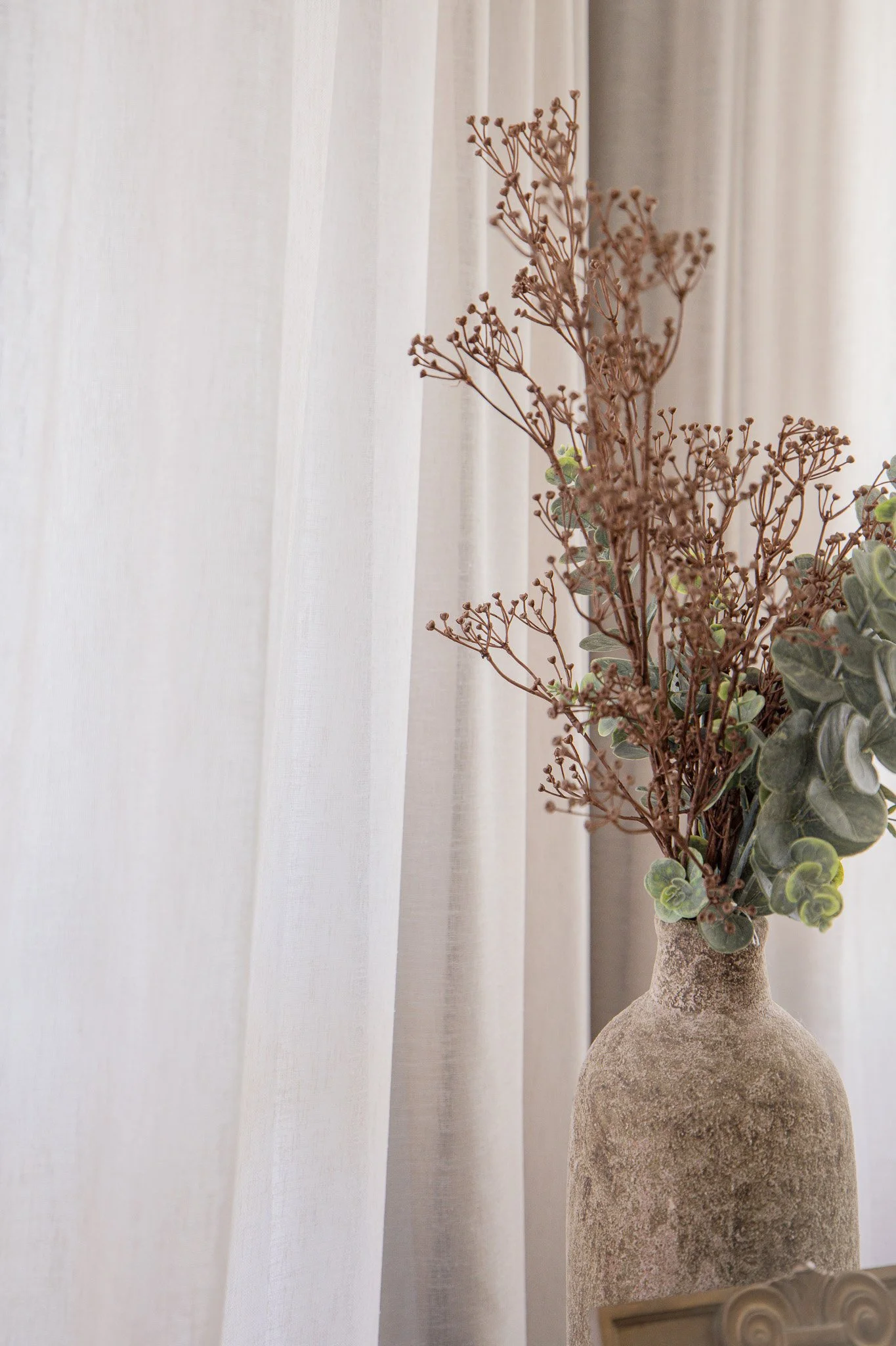 A beige textured vase filled with dried brown branches and green leaves in front of sheer white curtains.