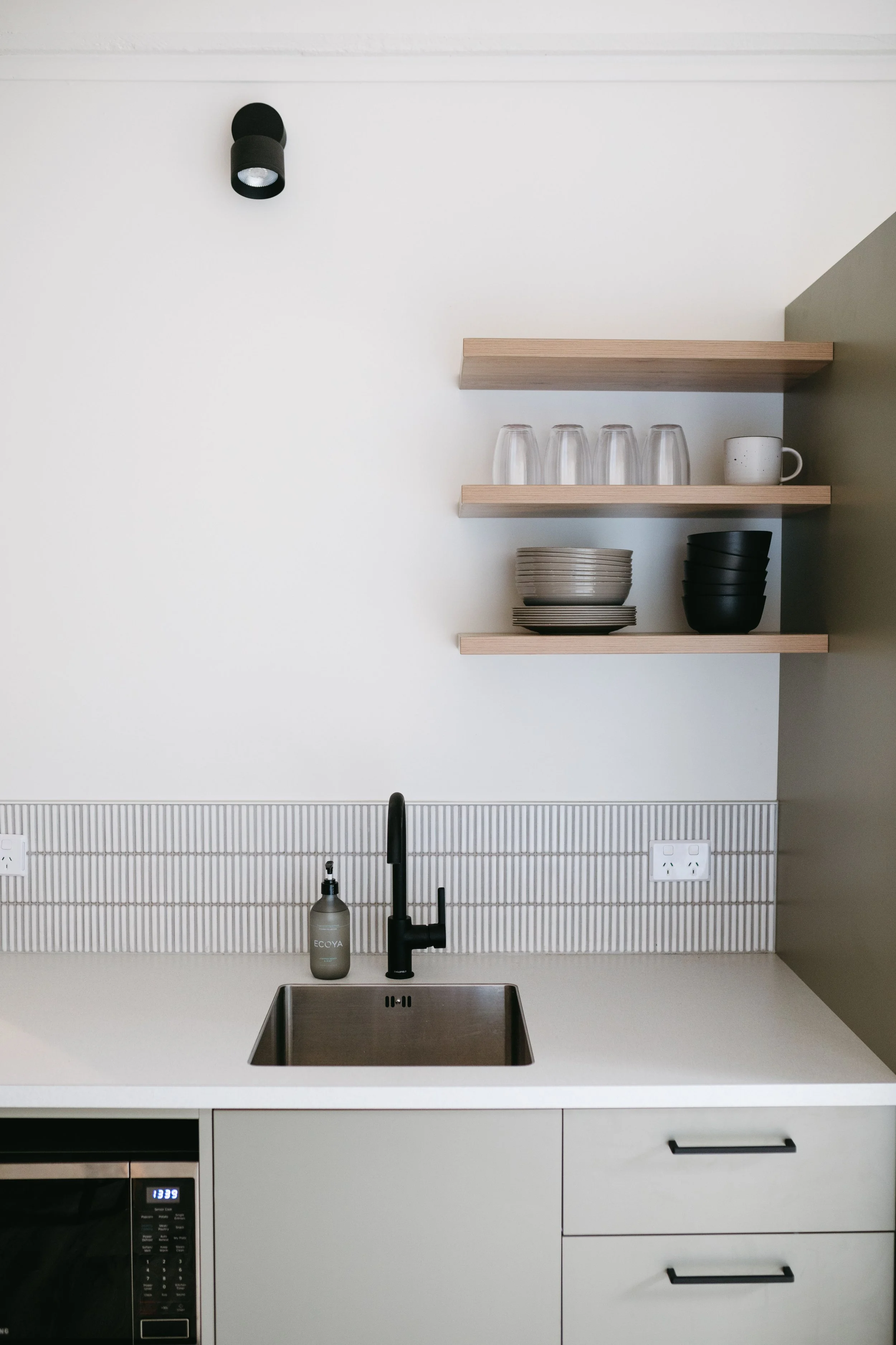 Modern kitchen with open wooden shelves, glassware, and bowls, a black faucet, soap dispenser, and a microwave below
