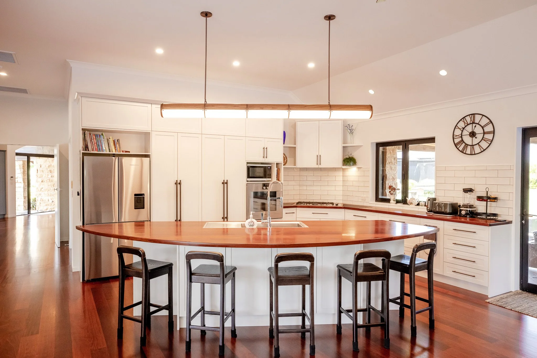 Modern kitchen with white cabinets, a large island with a wooden countertop, four black barstools, stainless steel refrigerator, built-in oven, microwave, and windowed black door.