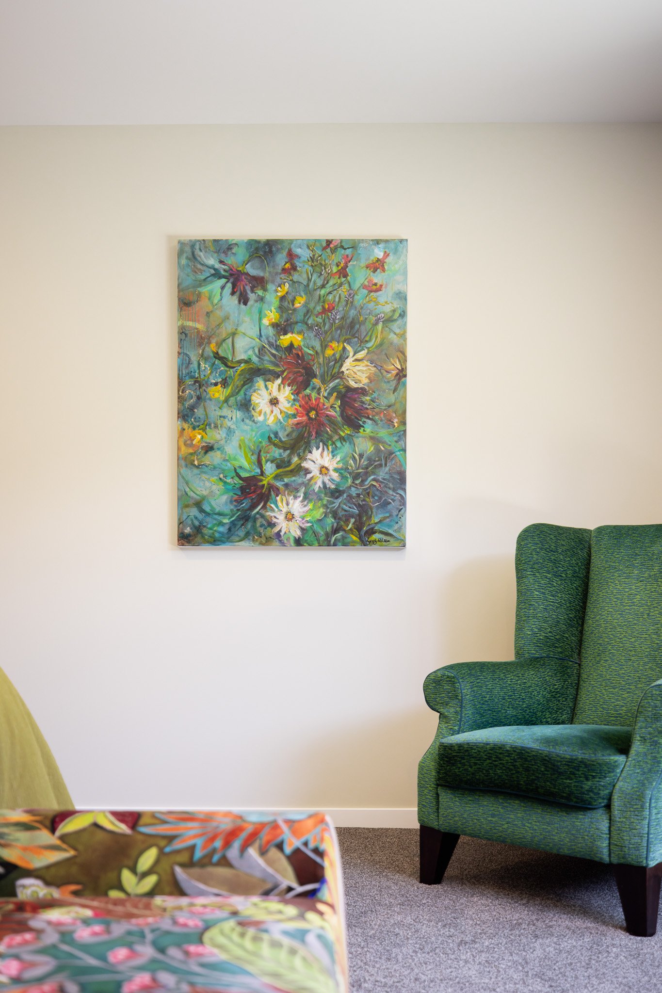 Interior of a room with a green armchair, a colorful floral painting on the white wall, and a part of a patterned sofa in the foreground.