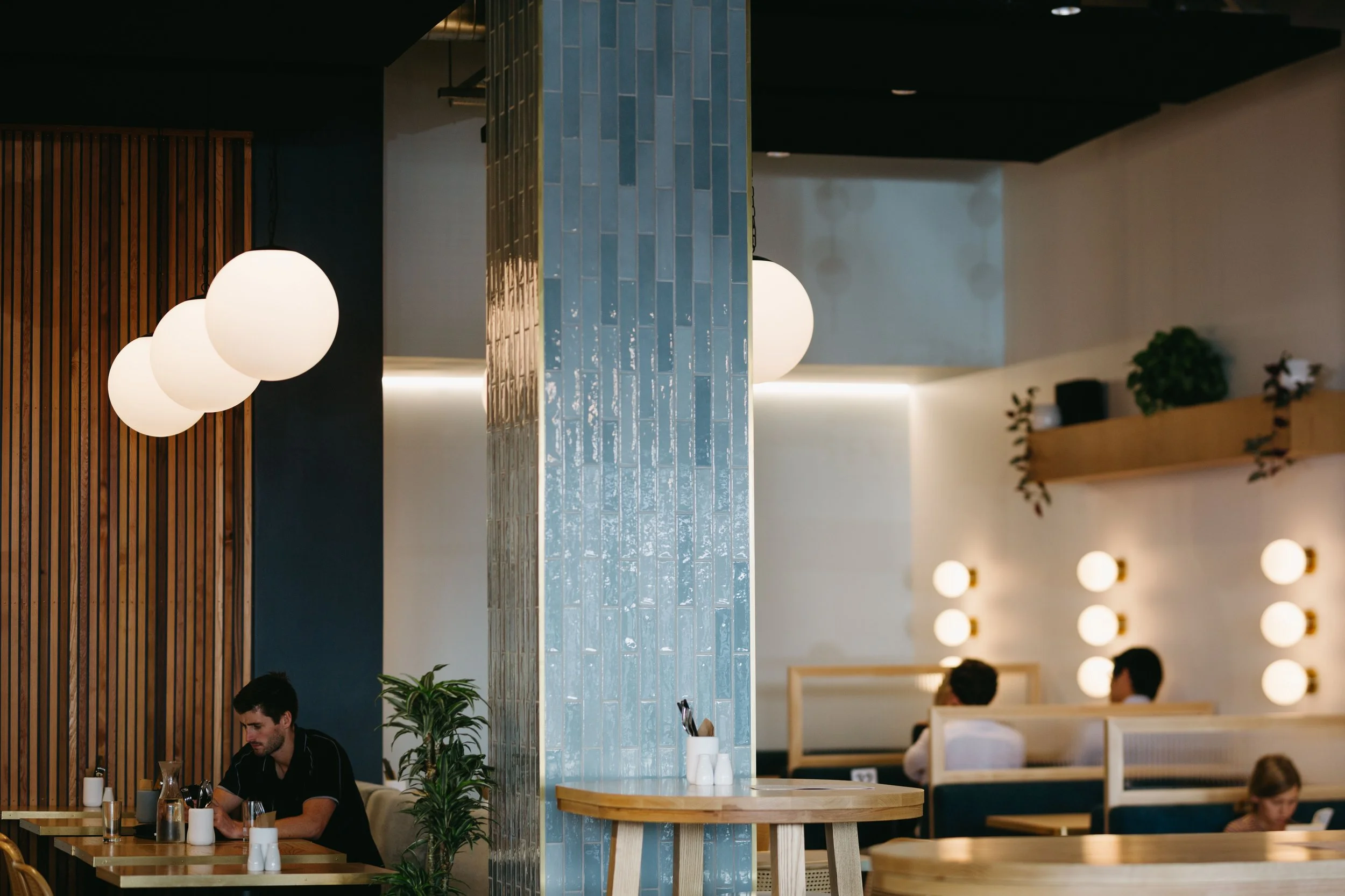 Interior of a modern restaurant with wooden accents, blue tiled pillar, spherical hanging lights, and booth seating with people dining.