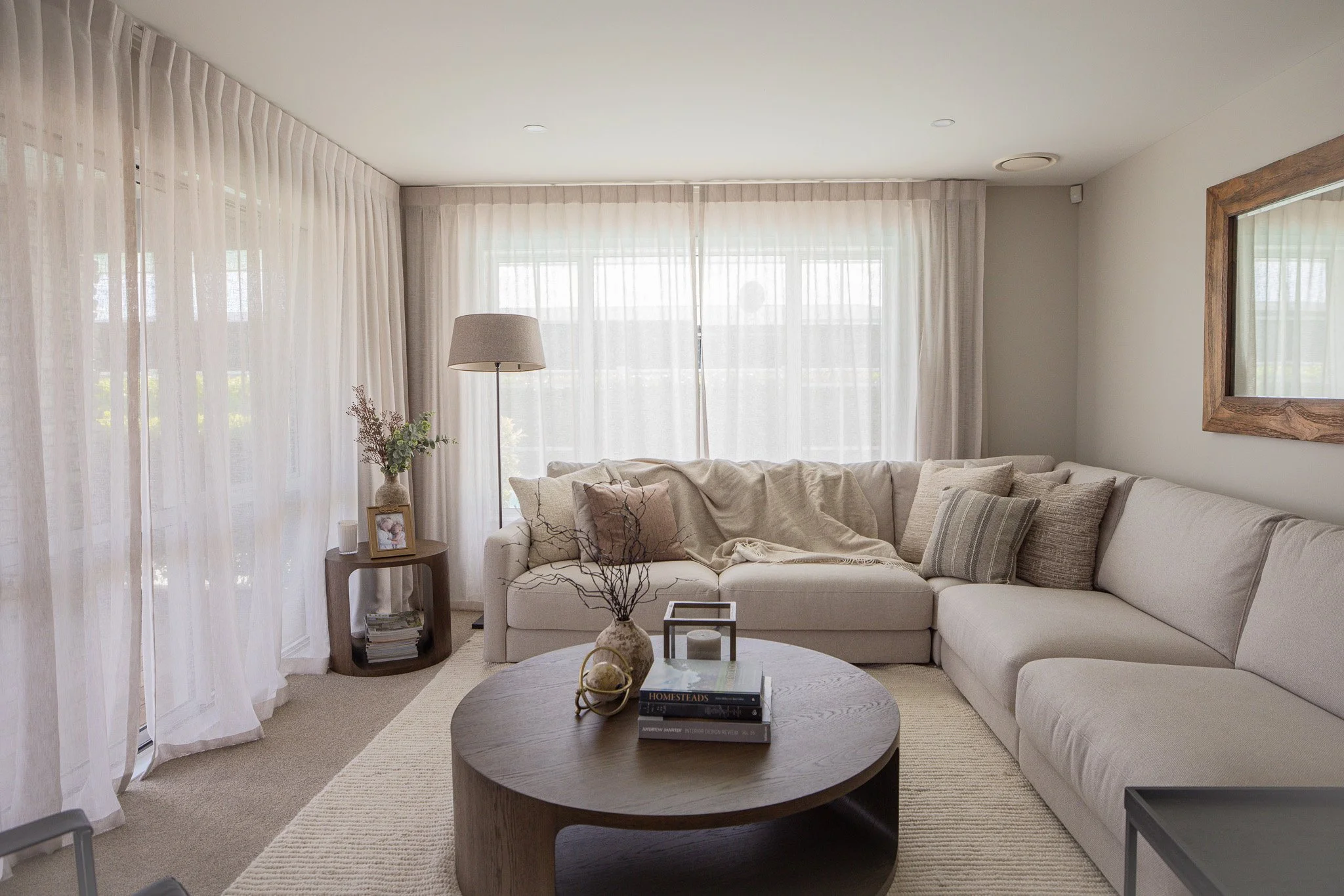 A bright living room with beige walls, sheer curtains, a sectional sofa with neutral pillows, a round wooden coffee table with books and decorative items, a side table with a photo frame and a vase, and a large mirror on the wall.