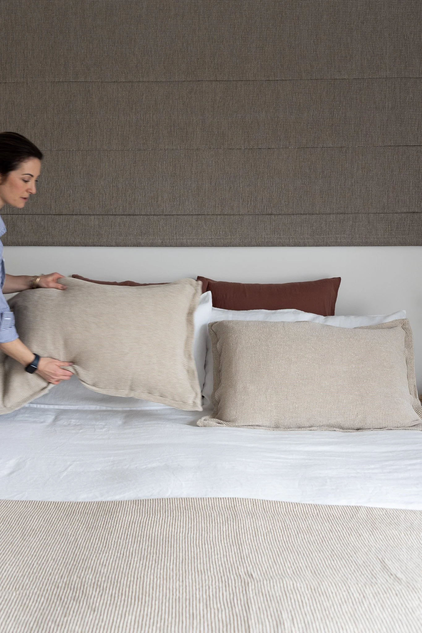 A woman arranging beige and brown pillows on a bed with white sheets and a beige bedspread.