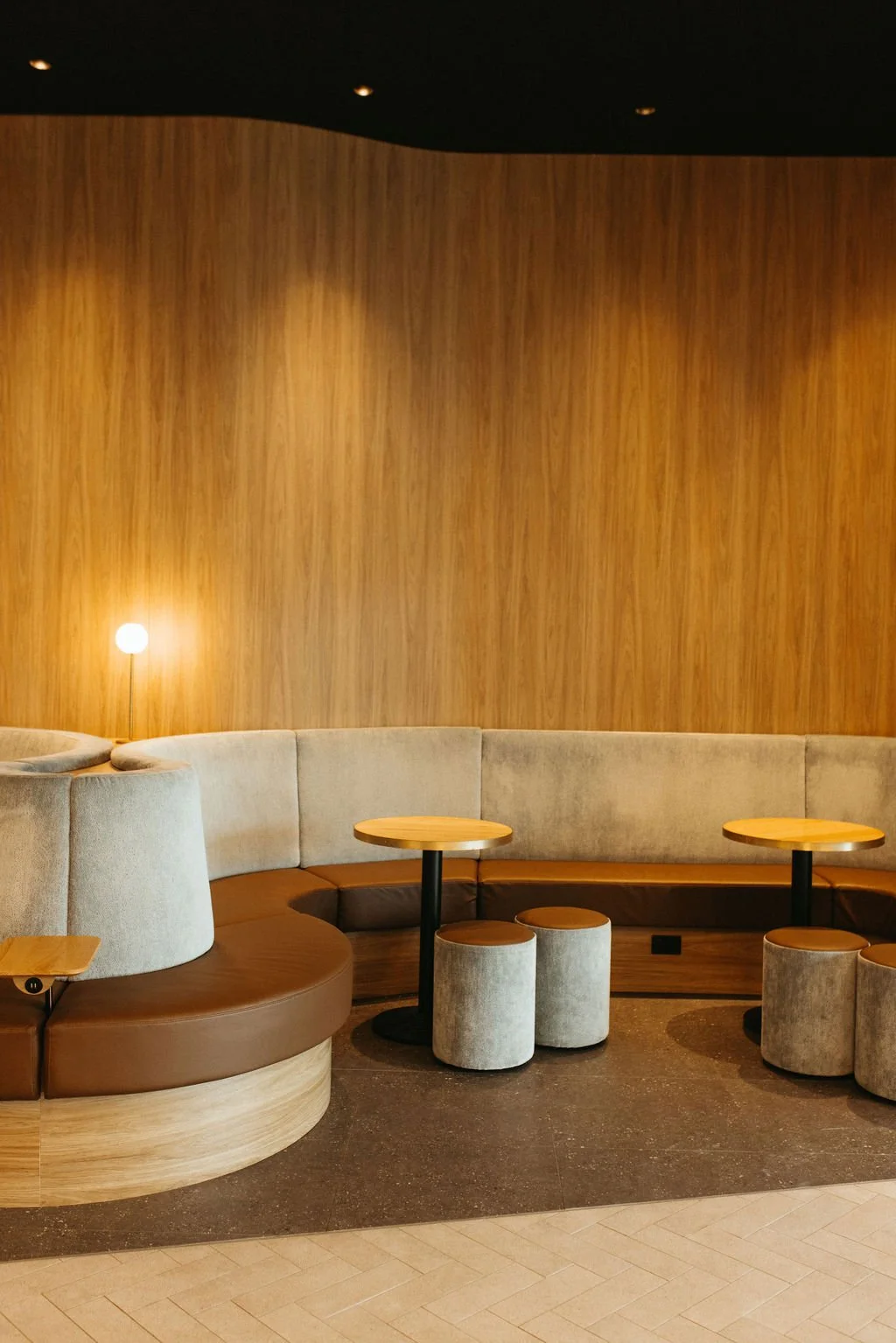A modern lounge area with curved beige and brown seating, small round tables, and a wooden wall background. There is a small lamp on the left side.
