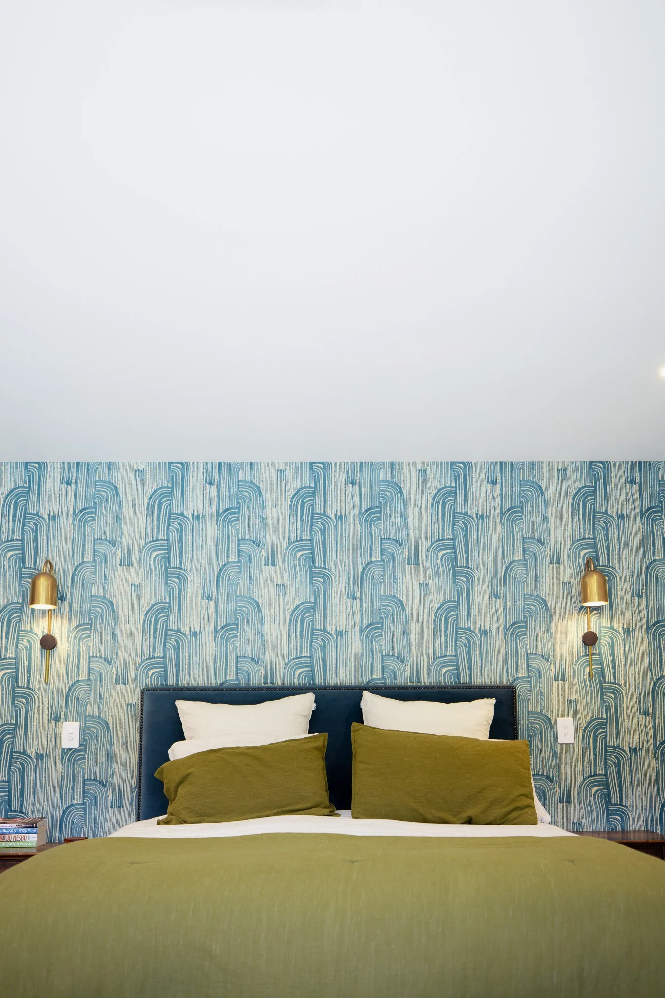 A neatly made bed with an olive green and white bedding set, positioned against a blue patterned accent wall with two gold wall-mounted reading lamps on either side.