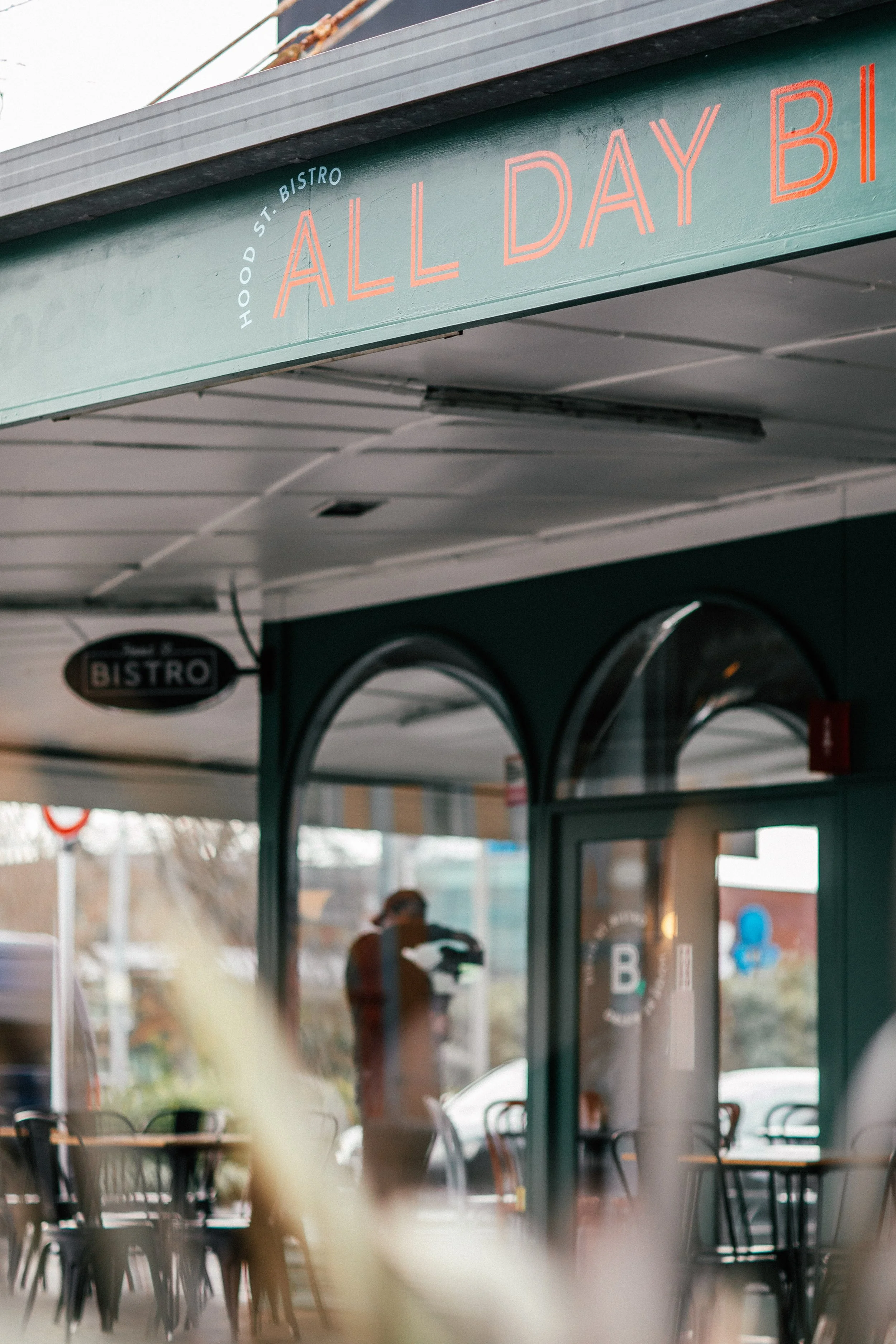 Exterior of a bistro restaurant named 'All Day Bist' on Hood Street, with outdoor seating and a person visible through the door.