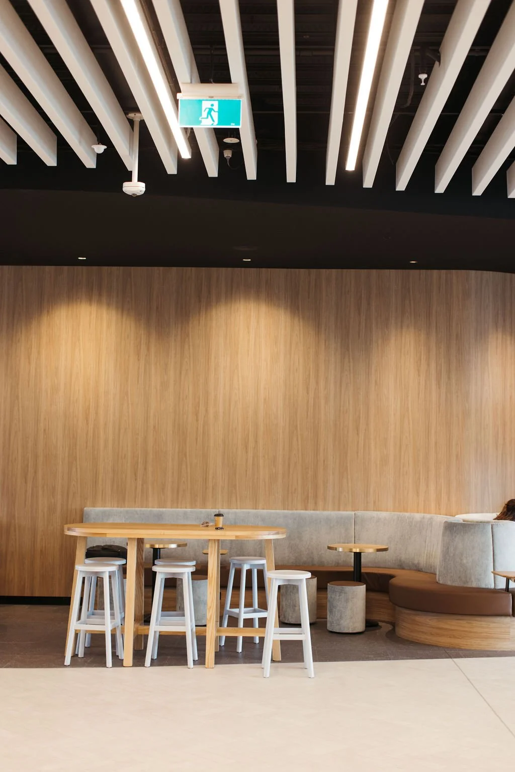 Empty modern lounge area with a curved beige and brown bench, small round tables, and bar stools in front of a wooden wall, with a ceiling featuring white slats and hanging light fixtures, and an exit sign hanging from the ceiling.