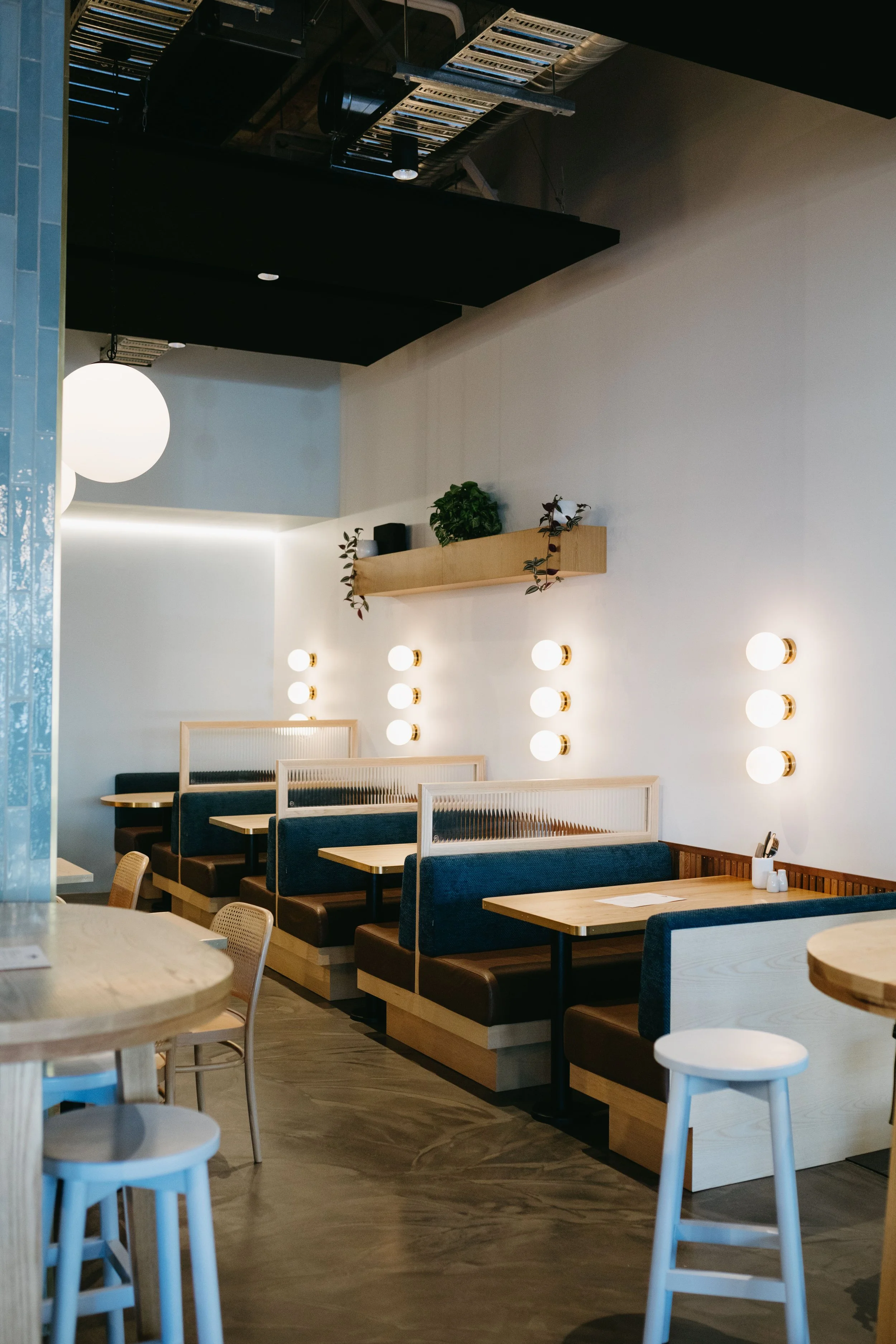 Empty modern restaurant with wooden booths, white walls, and minimal decor, including a floating shelf with plants and multiple wall-mounted light fixtures.