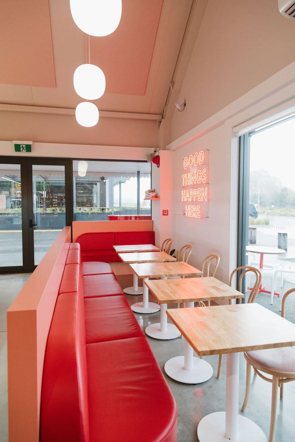 Interior of a cafe with red seating, wooden tables, white chairs, and a neon sign that reads 'Good Things Happen Here' on the wall. Large windows allow natural light to illuminate the space.