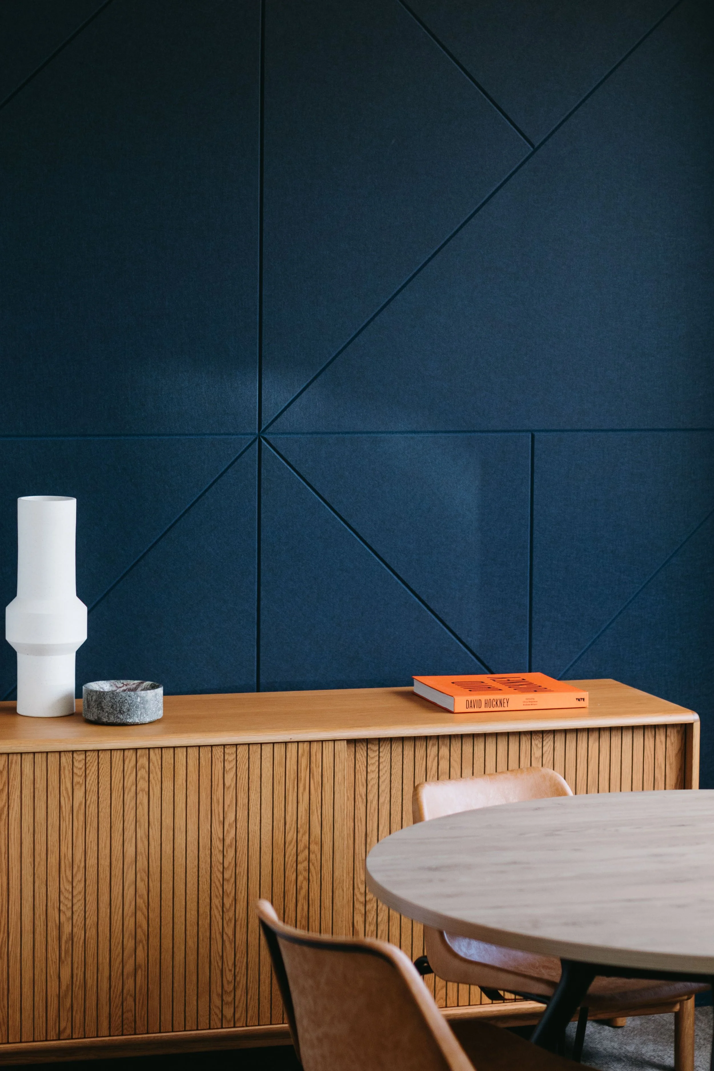 A modern interior with a wood sideboard, a tall white vase, a round gray stone object, and an orange book on the sideboard, with a dark blue geometric wall and a round wooden table in the foreground.