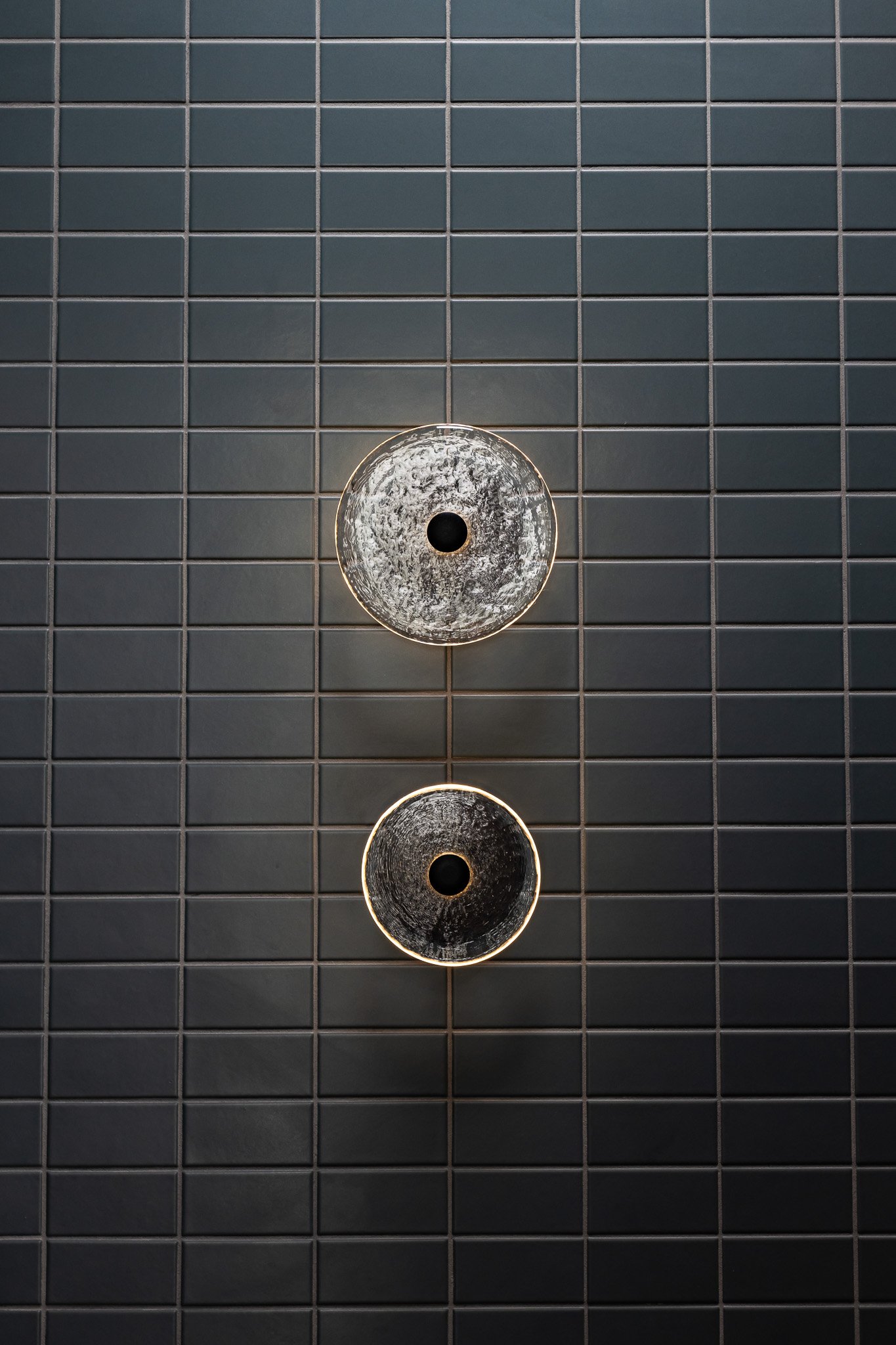 Two round ceiling lights with a textured surface, mounted on a wall with dark gray square tiles arranged in a grid pattern.
