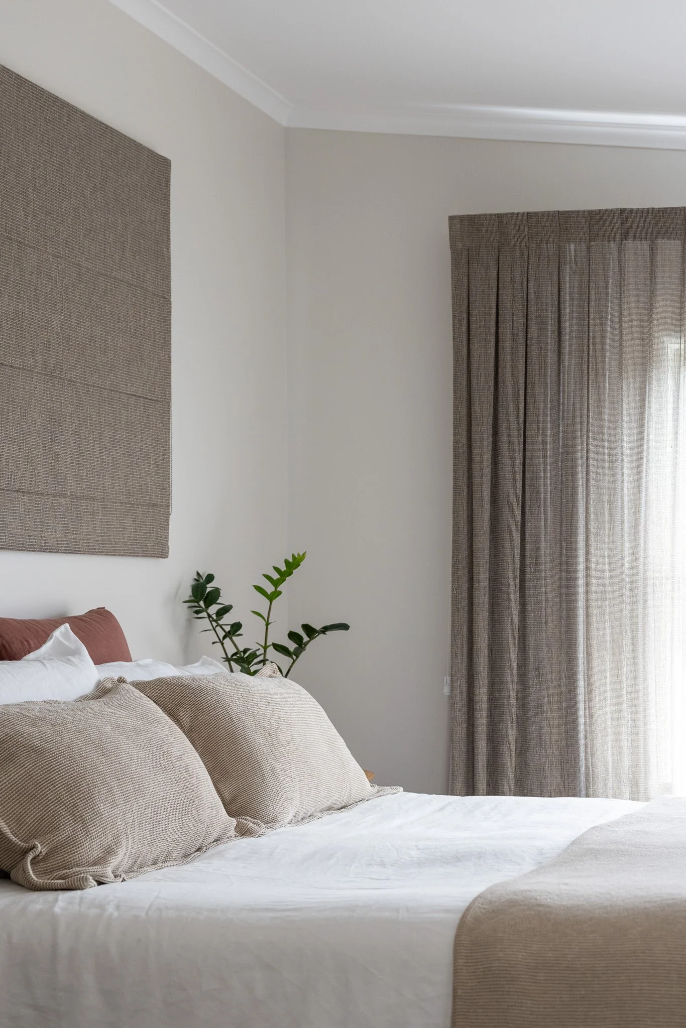 A neatly made bed with beige pillows, white bedding, and a brown headboard in a minimalist bedroom with neutral-colored walls, a potted plant, and a window with brown curtains.