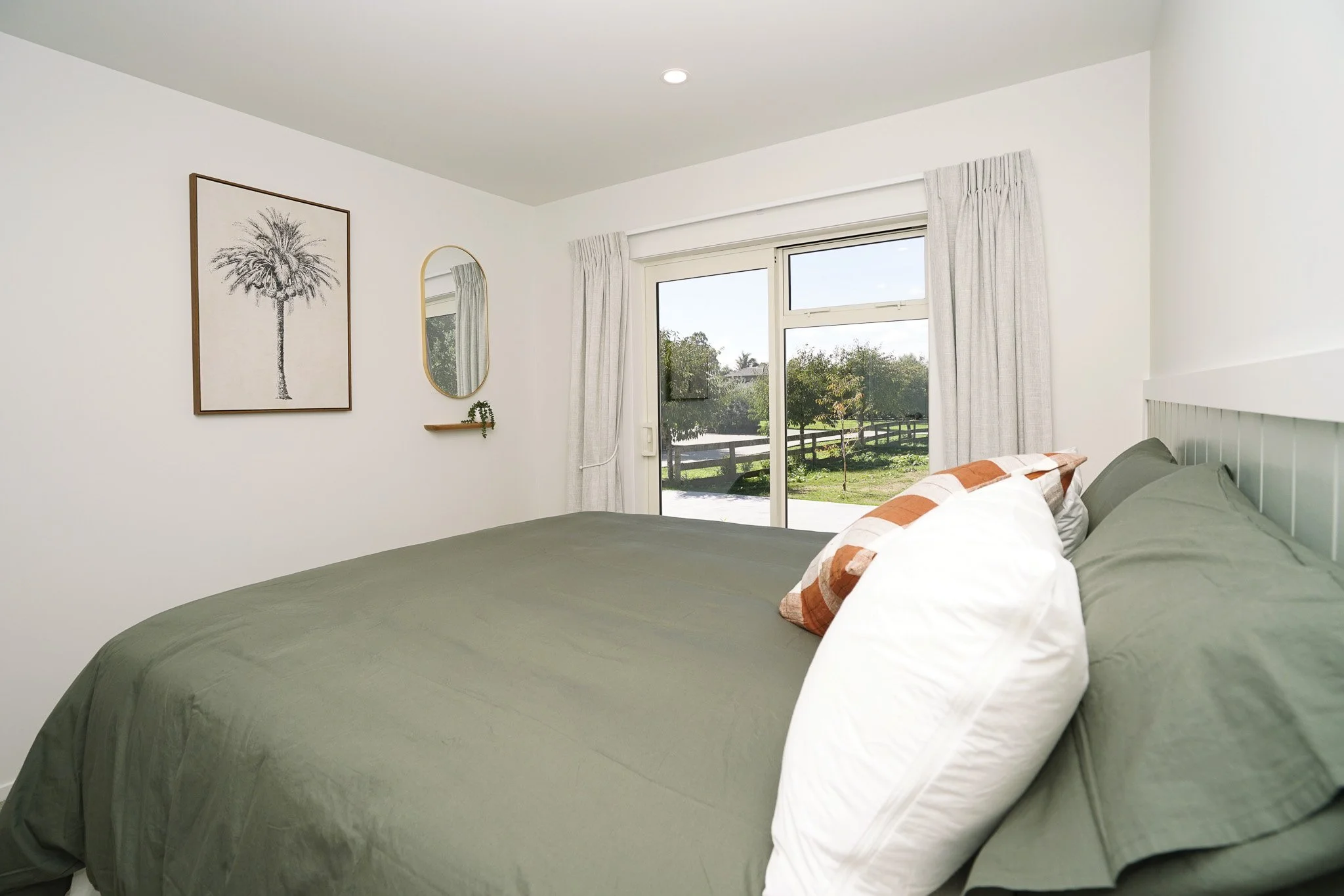 Bedroom with a large bed covered with green bedding and pillows, window with white curtains, wall decor including a palm tree artwork and a mirror, and a view of greenery outside.