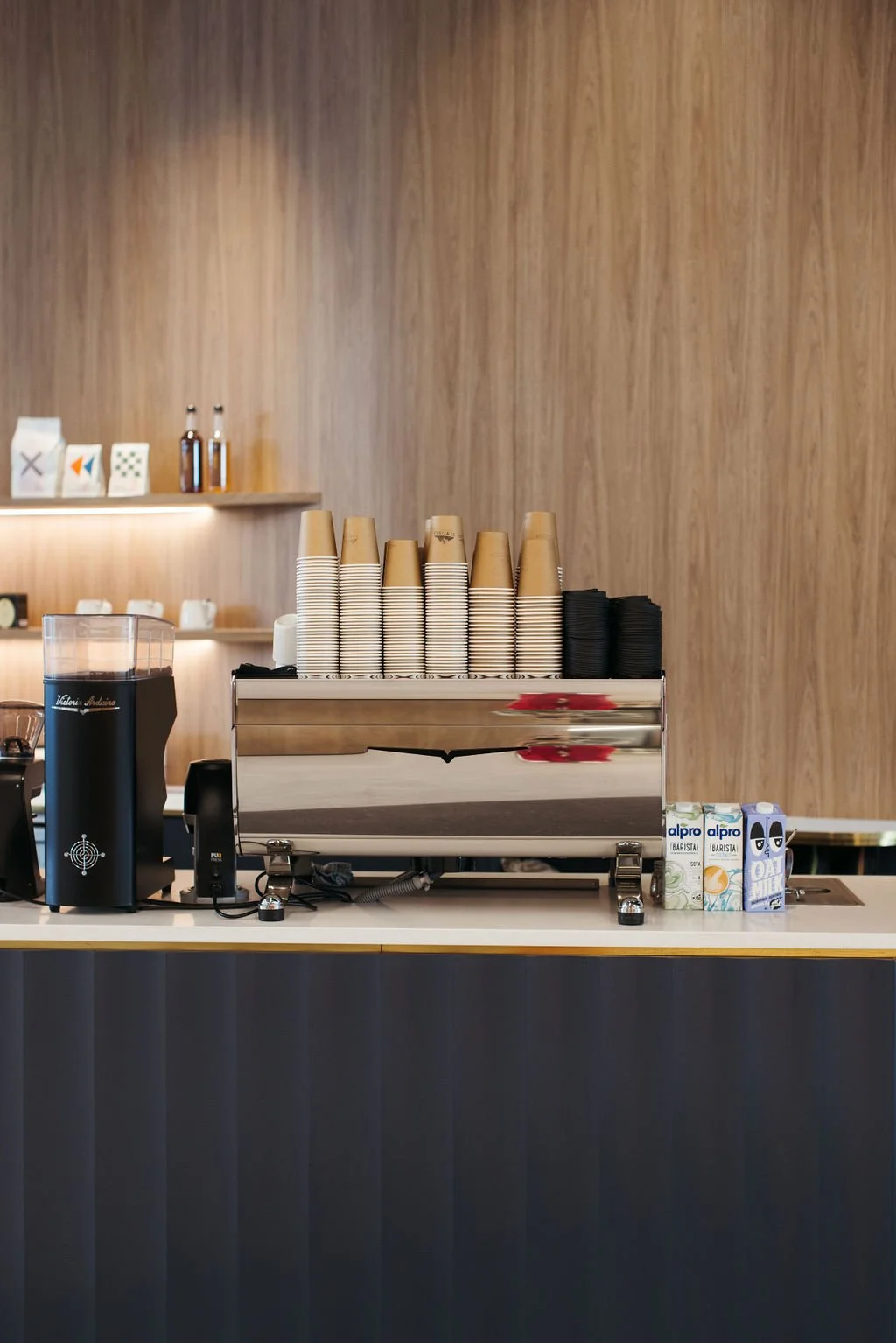 Coffee station with disposable cups, coffee machine, and milk cartons on a bar counter in a cafe or coffee shop.