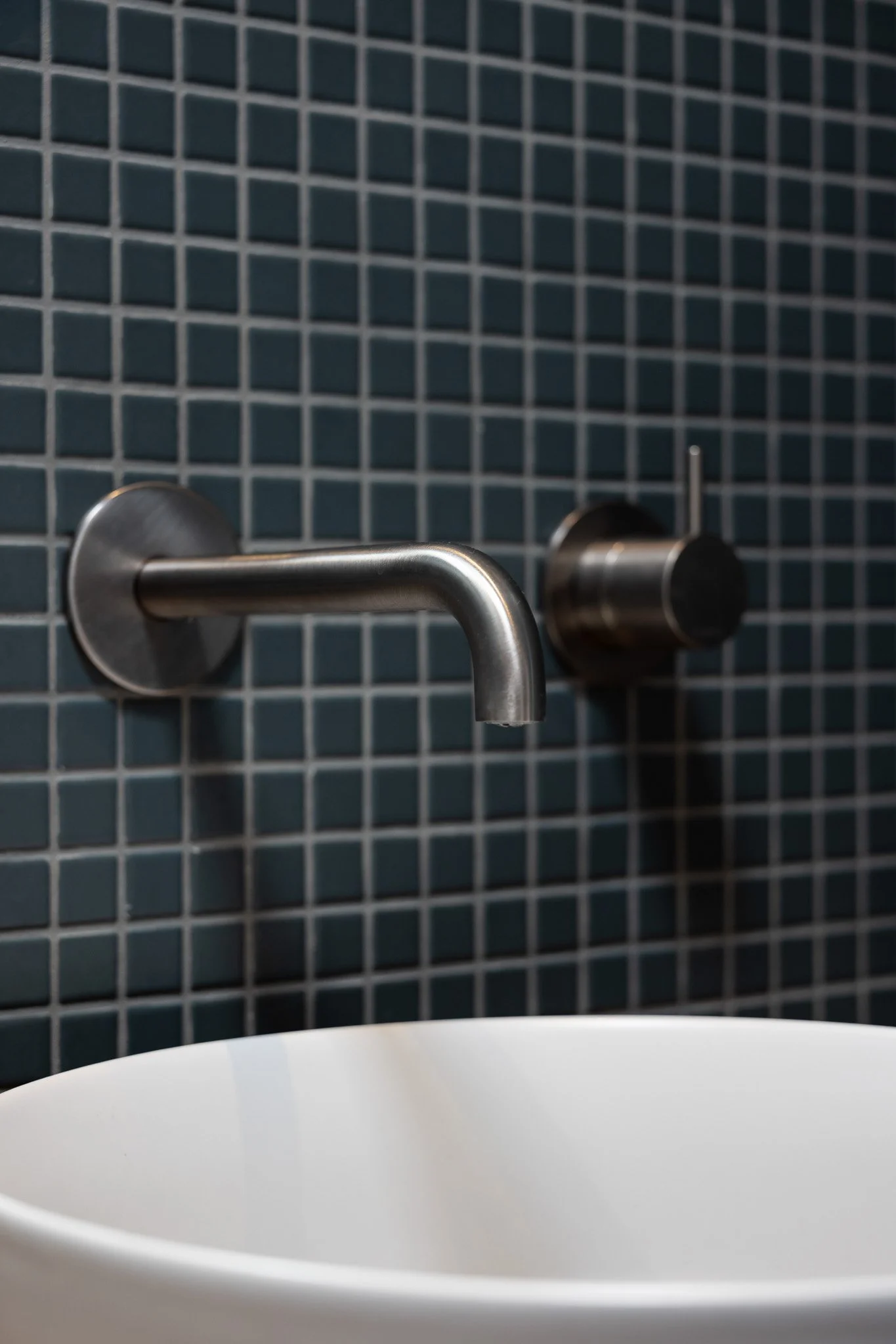 Close-up of a metallic water faucet on a tiled wall in a bathroom with a partially visible white sink below.