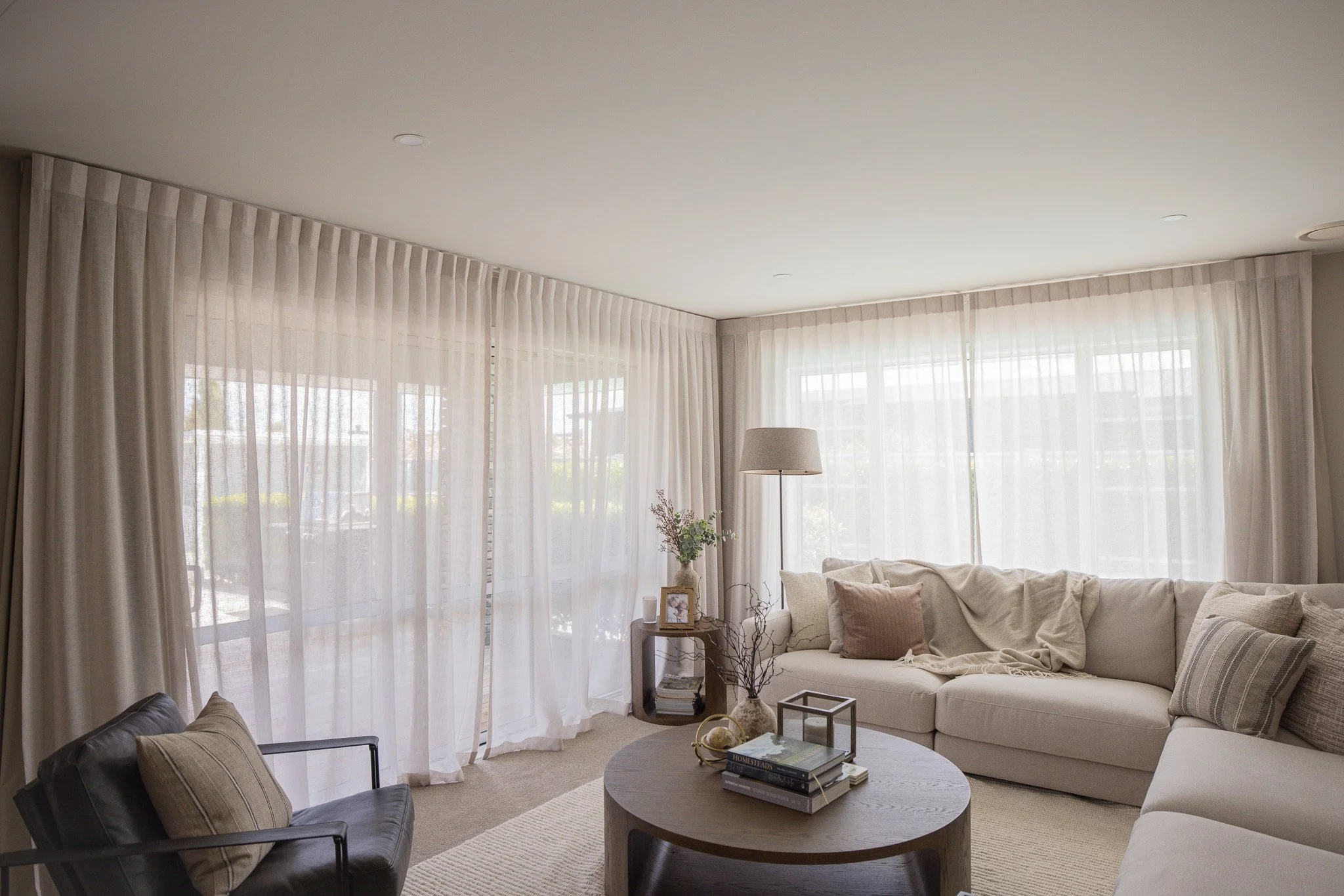 Living room with beige sectional sofa, black armchair, round coffee table, and sheer white curtains covering large windows.