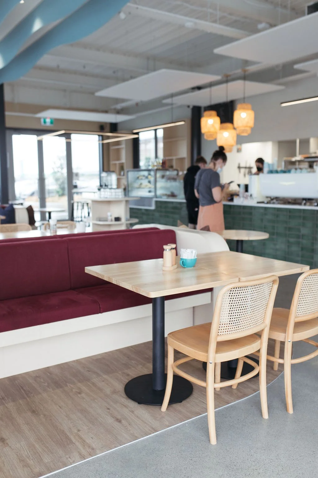 Interior of a modern café with wooden tables, beige chairs, a seating bench with maroon cushions, and a counter with a few customers ordering. The café has hanging pendant lights and large windows letting in natural light.