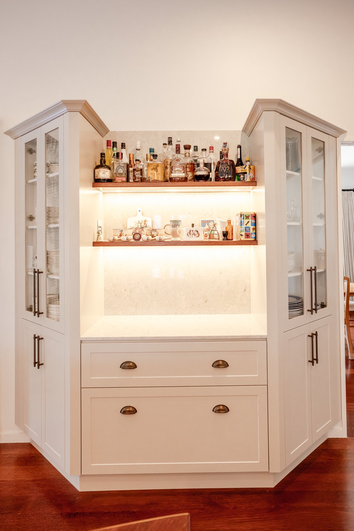 A white built-in cabinet with glass-paneled doors, brass handles, and two drawers, housing a collection of bottles, dishes, and decorative items, illuminated by warm lighting.