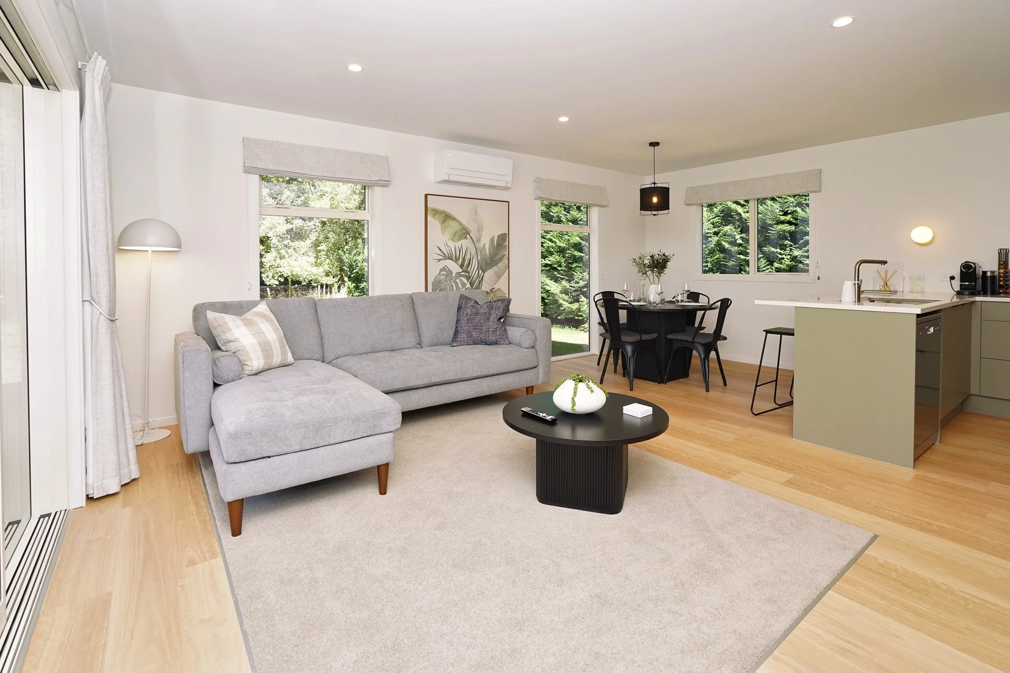 Open-concept living room and kitchen with light wood flooring, gray sectional sofa, black round coffee table, dining table with four chairs, and large windows showing greenery outside.