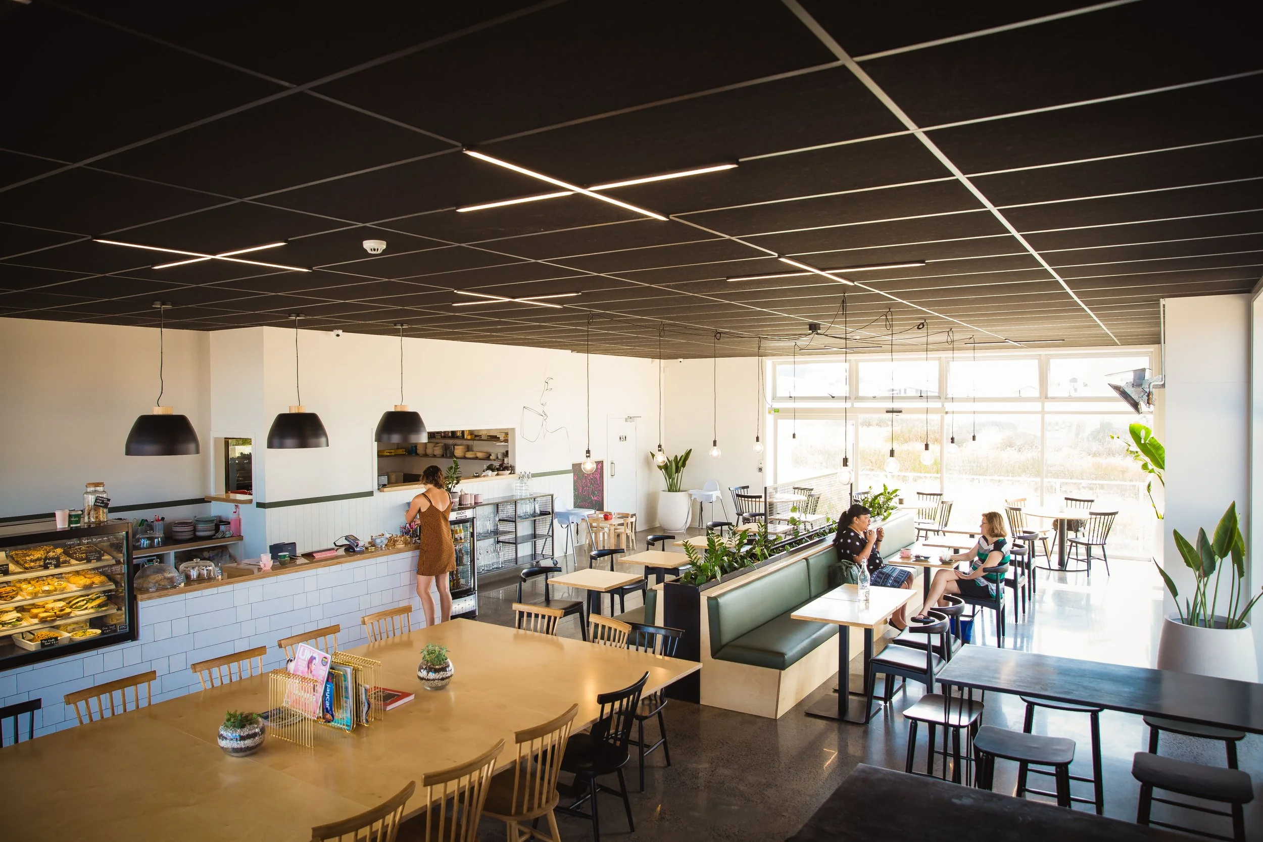 Inside a modern cafe with a black ceiling, hanging pendant lights, and large windows letting in natural light. There are various tables and chairs, with two women seated and talking near the middle. A person behind the counter prepares food, and ther
