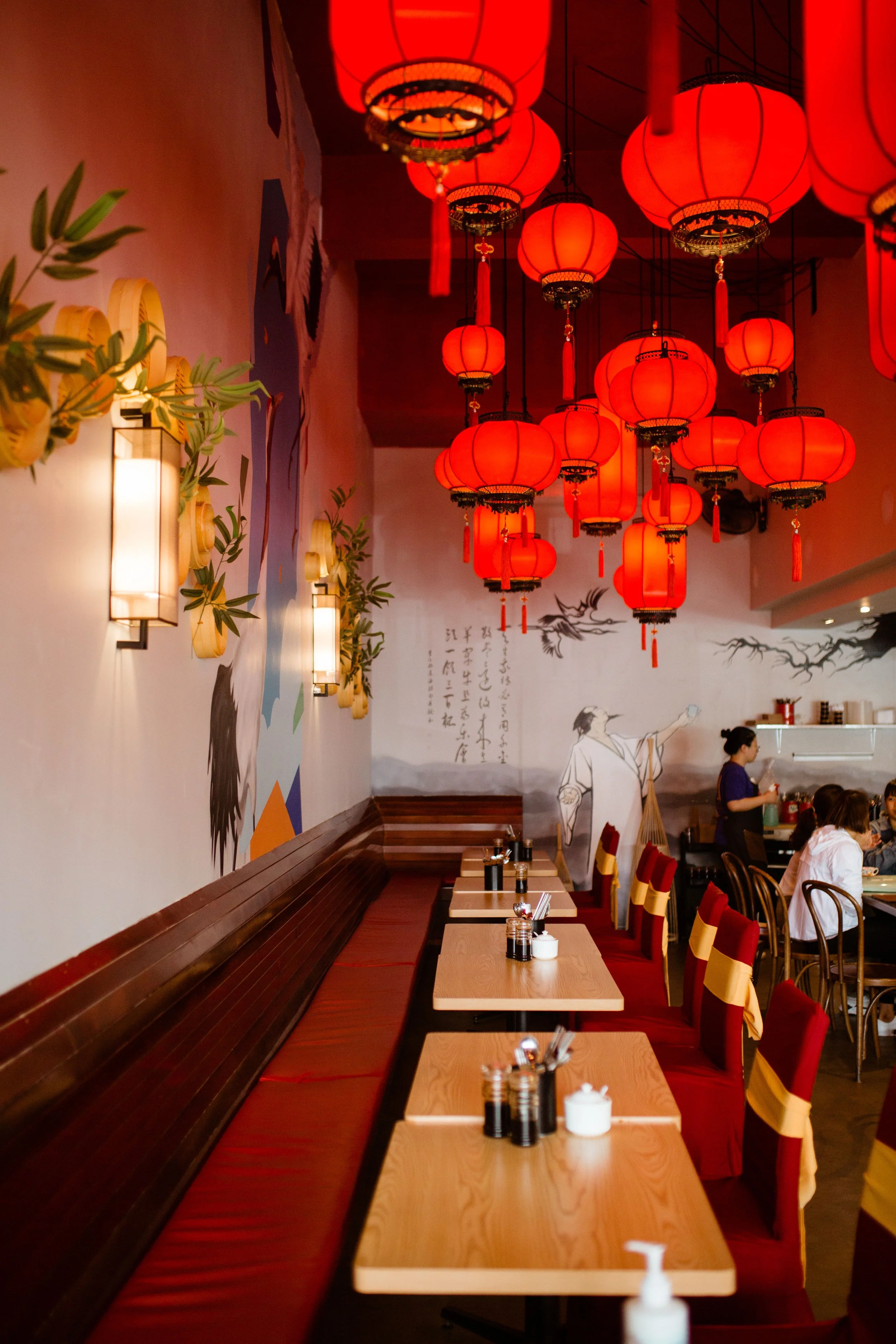 Interior of a restaurant decorated with numerous red Chinese lanterns hanging from the ceiling, a wall with traditional Chinese artwork and calligraphy, and tables set with condiments and utensils.