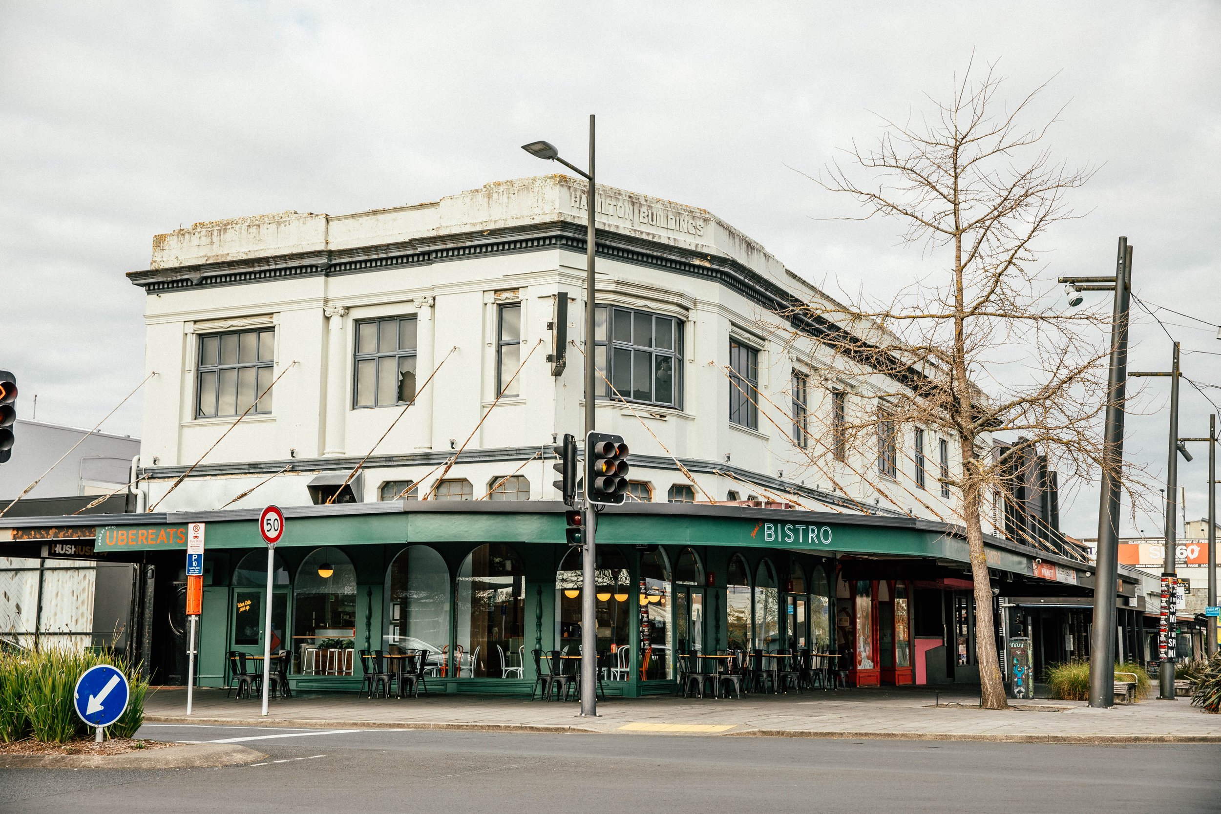 Corner building with white upper stories and green ground level with a sign labeled "BISTRO." Empty outdoor seating area with black chairs and tables. Tree with no leaves in front. Traffic light and street signs at corner.