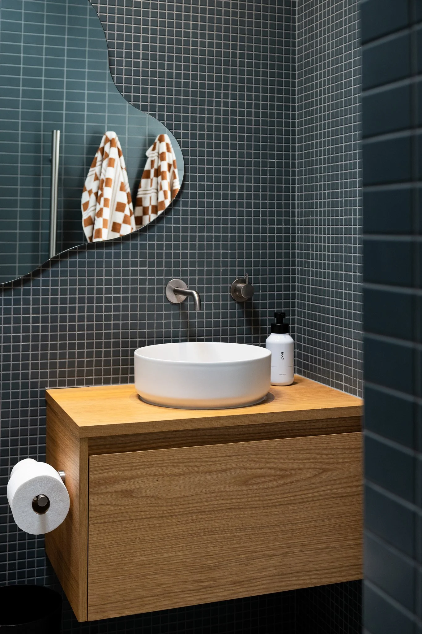 Modern bathroom with dark mosaic tile wall, round white vessel sink on wooden vanity, wall-mounted faucet, bottle of soap, mirror, and two plaid towels hanging on a pole.