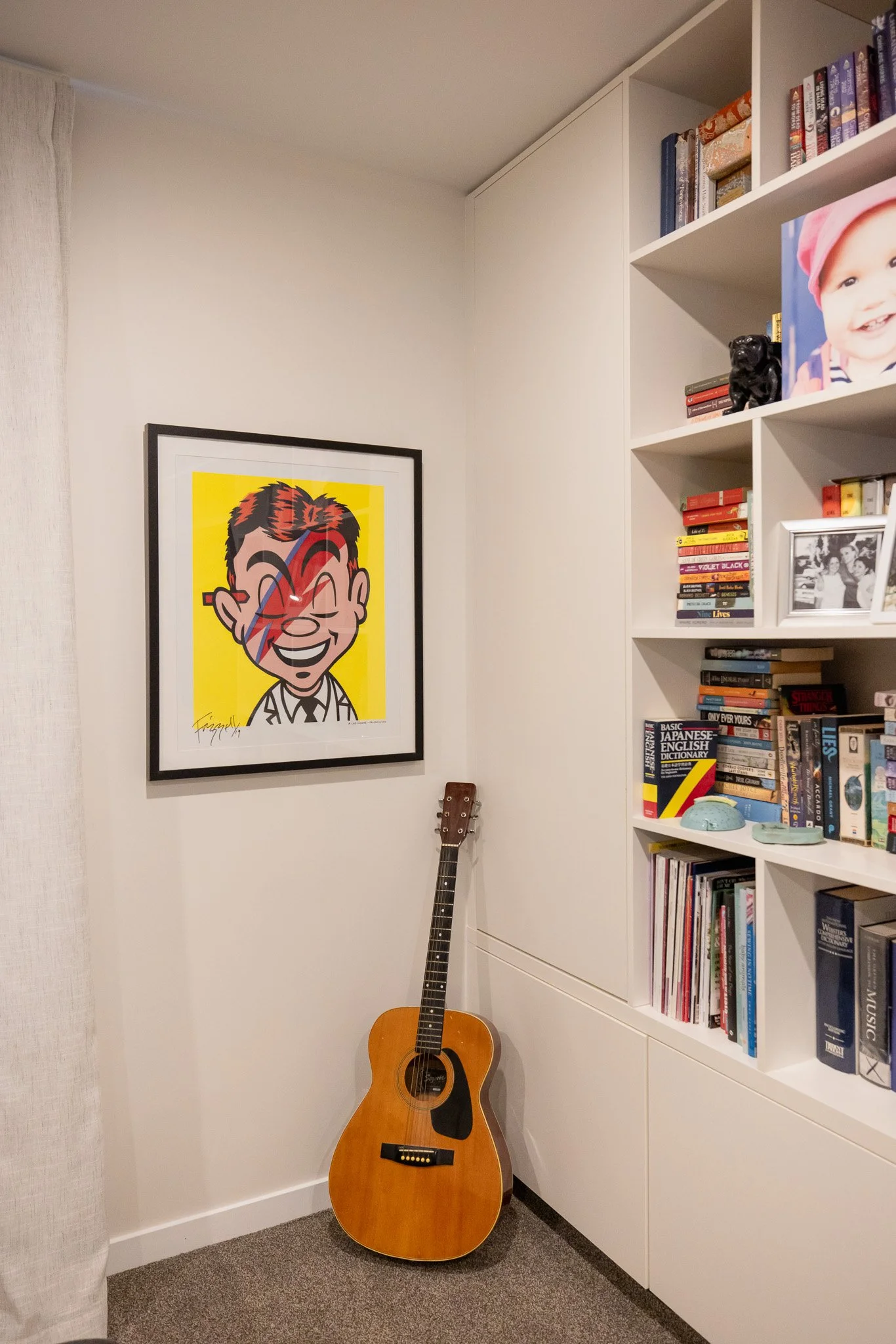 A corner of a room with a guitar leaning against the white wall near a bookshelf filled with books, photos, and decorative items. A framed pop art portrait of a smiling man with a lightning bolt across his face hangs on the wall.
