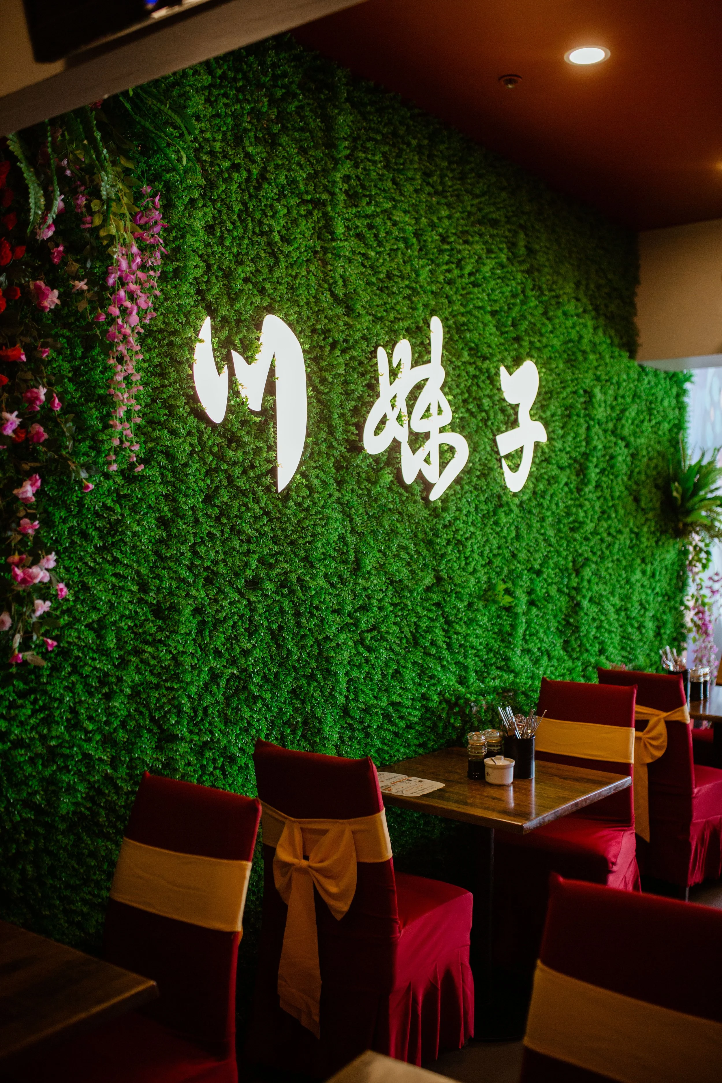A restaurant interior with a green faux plant wall with white illuminated Chinese characters, red velvet chairs with yellow sashes, and wooden tables with condiments and utensils.