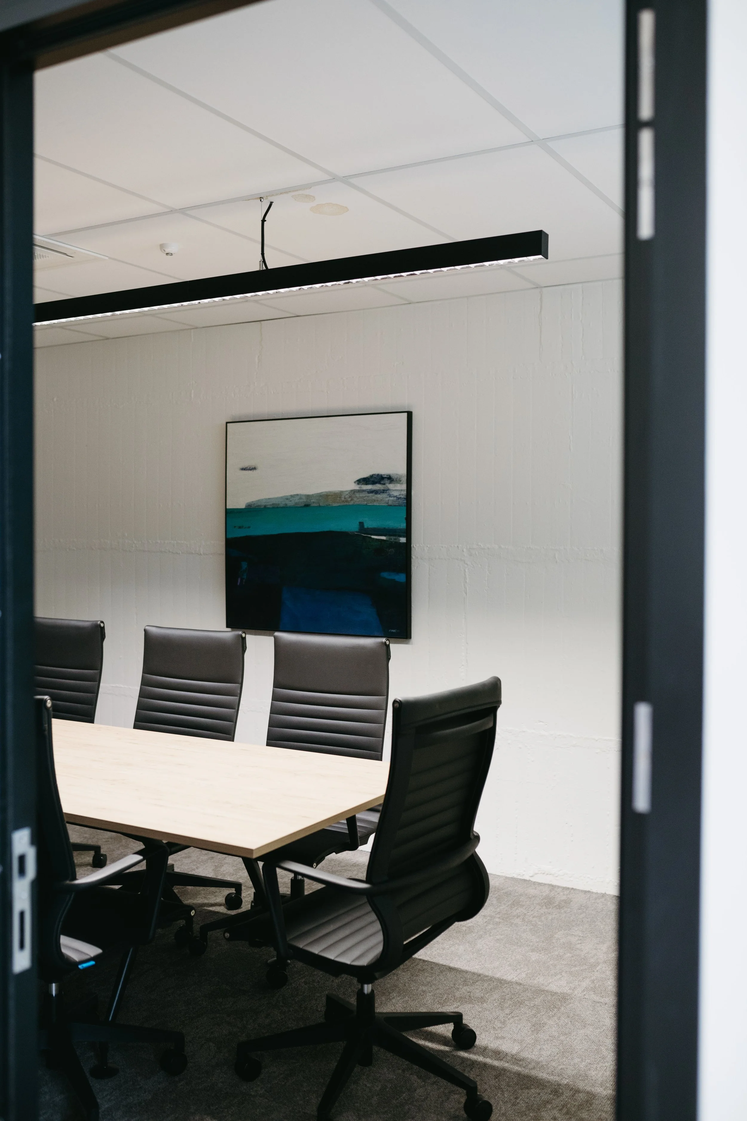 Empty conference room with six black leather office chairs around a light wooden table, a framed landscape painting on the white wall, and a modern linear ceiling light fixture.