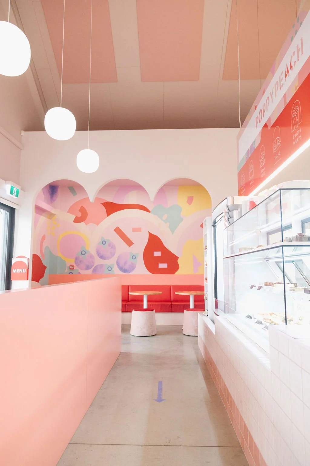 Inside a pastel pink and white ice cream shop with abstract wall art, white ceiling, hanging globular lights, and a pink seating area with red cushioned benches and small tables. Ice cream display case on the right.