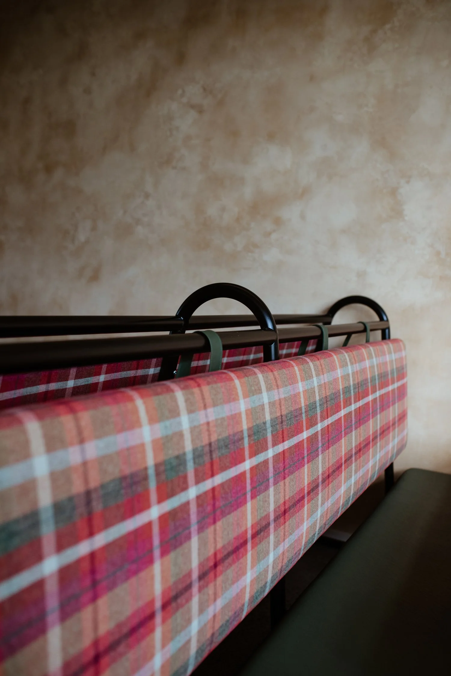 Close-up of a red plaid upholstered bench or seat with metal and plastic hooks or bars behind it, against a textured beige wall.
