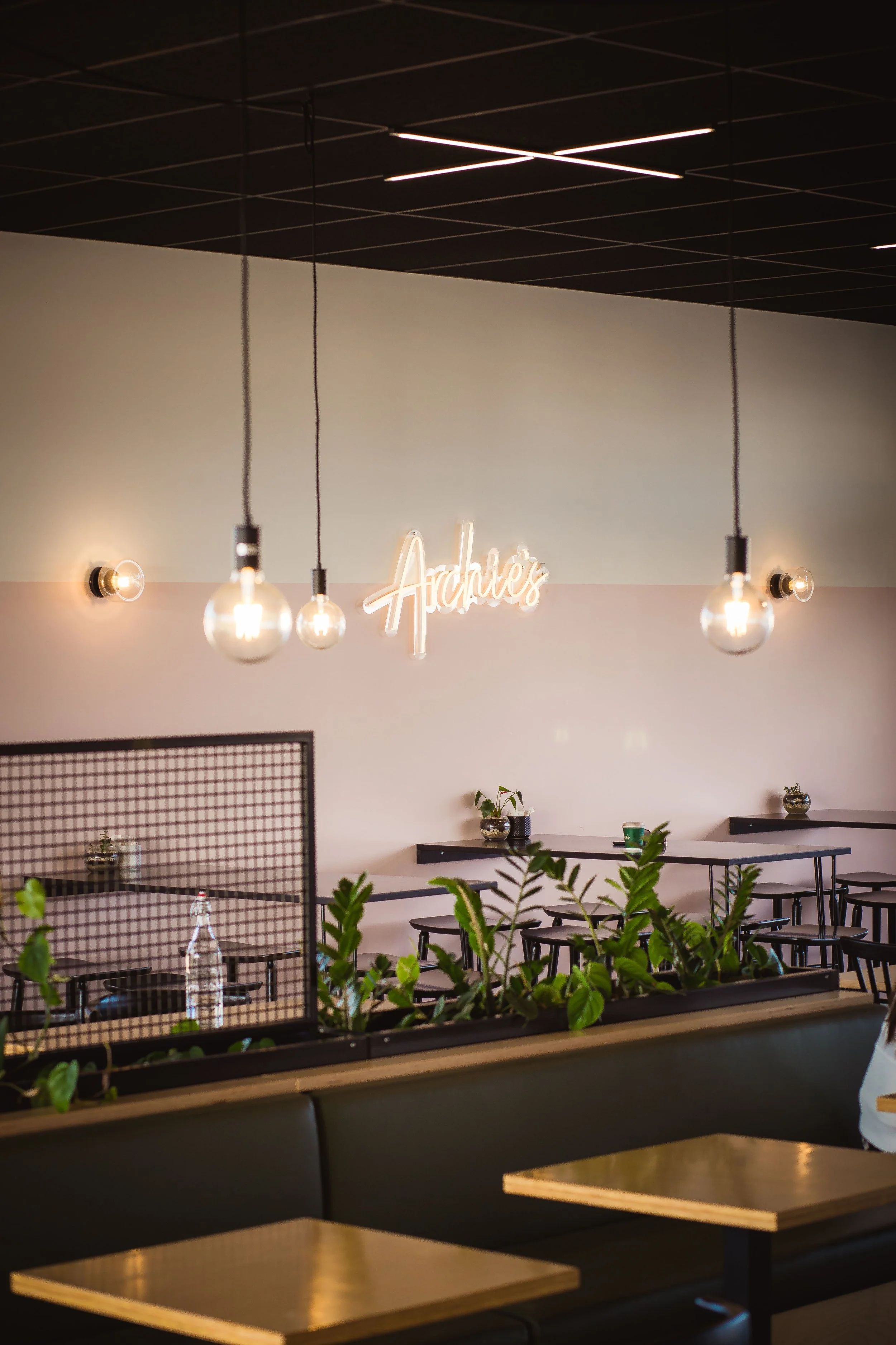 Interior of a modern coffee shop or restaurant with hanging light bulbs, a neon sign reading 'Archives' on the wall, and a mix of tables and seating areas with plants and decorative items.