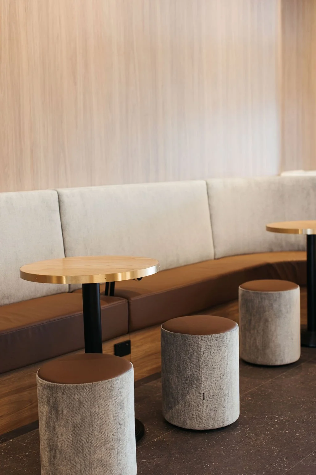 Modern lobby seating area with beige upholstered bench, three round ottomans, and two small round tables with wooden tops and black bases.