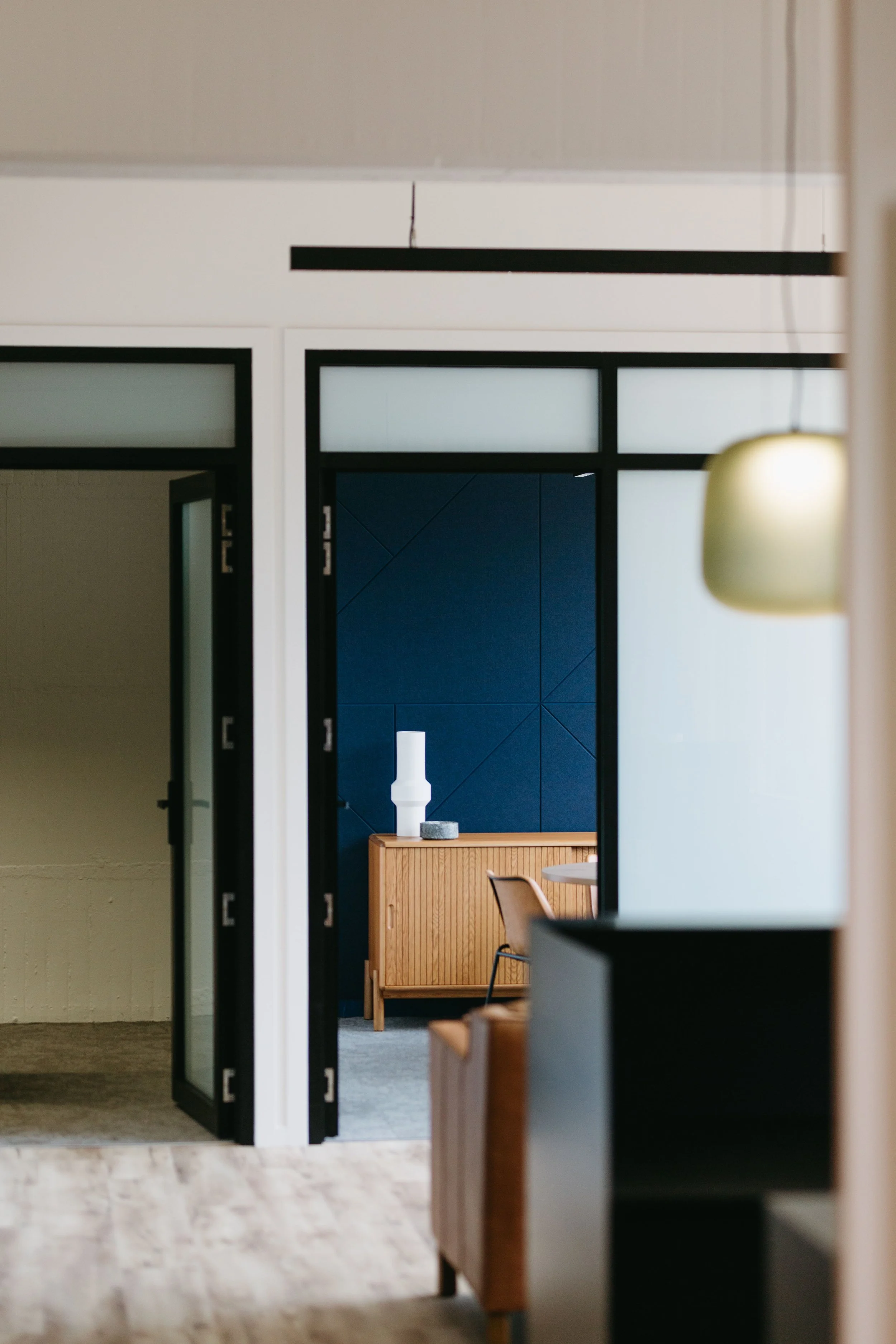 Interior view of a modern room with a wooden sideboard, blue wall panels, and a white sculptural vase on top, seen through an open doorway.