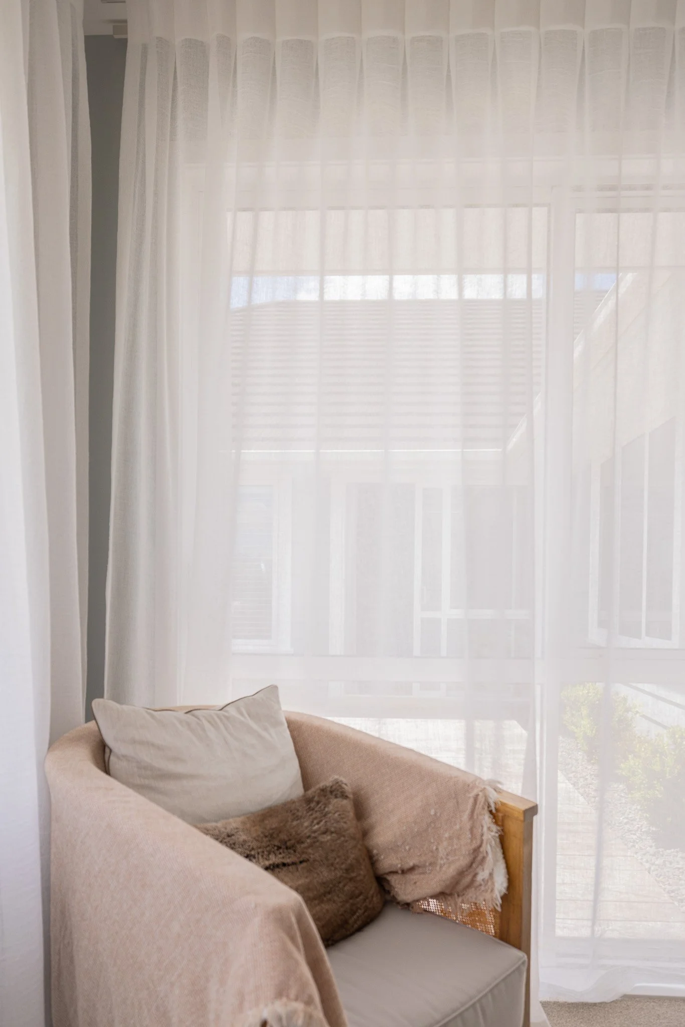 A cozy corner with a beige armchair, decorated with a white pillow, a brown furry pillow, and a beige throw blanket, next to large windows with sheer white curtains letting in natural light.