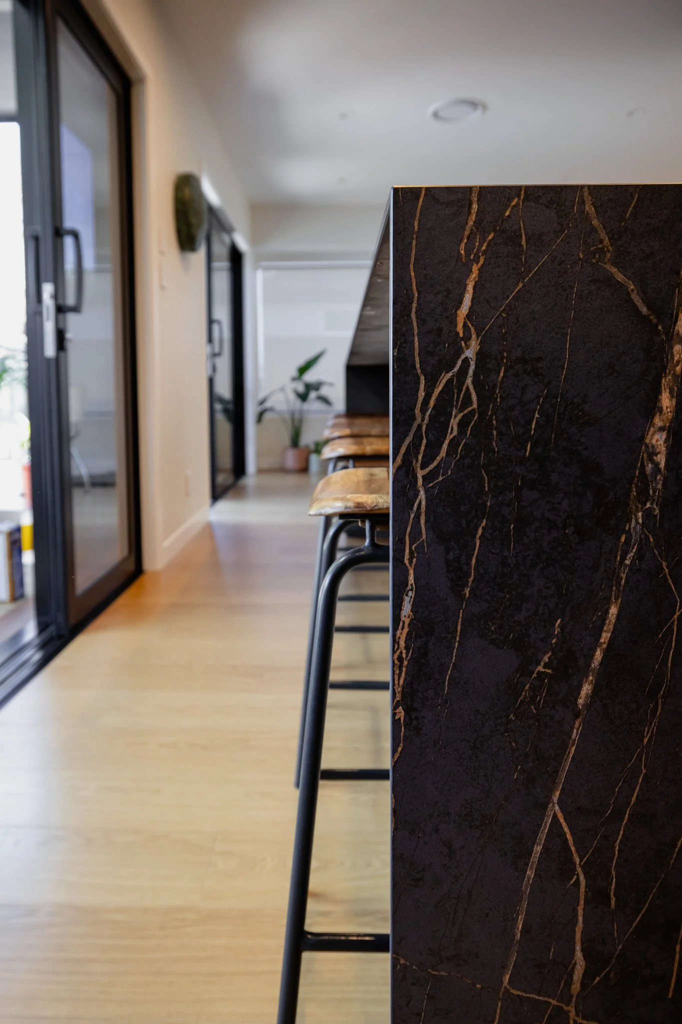 Close-up of a black marble kitchen island with gold veining, stainless steel and wooden bar stools, and a modern interior with sliding glass doors and a potted plant.