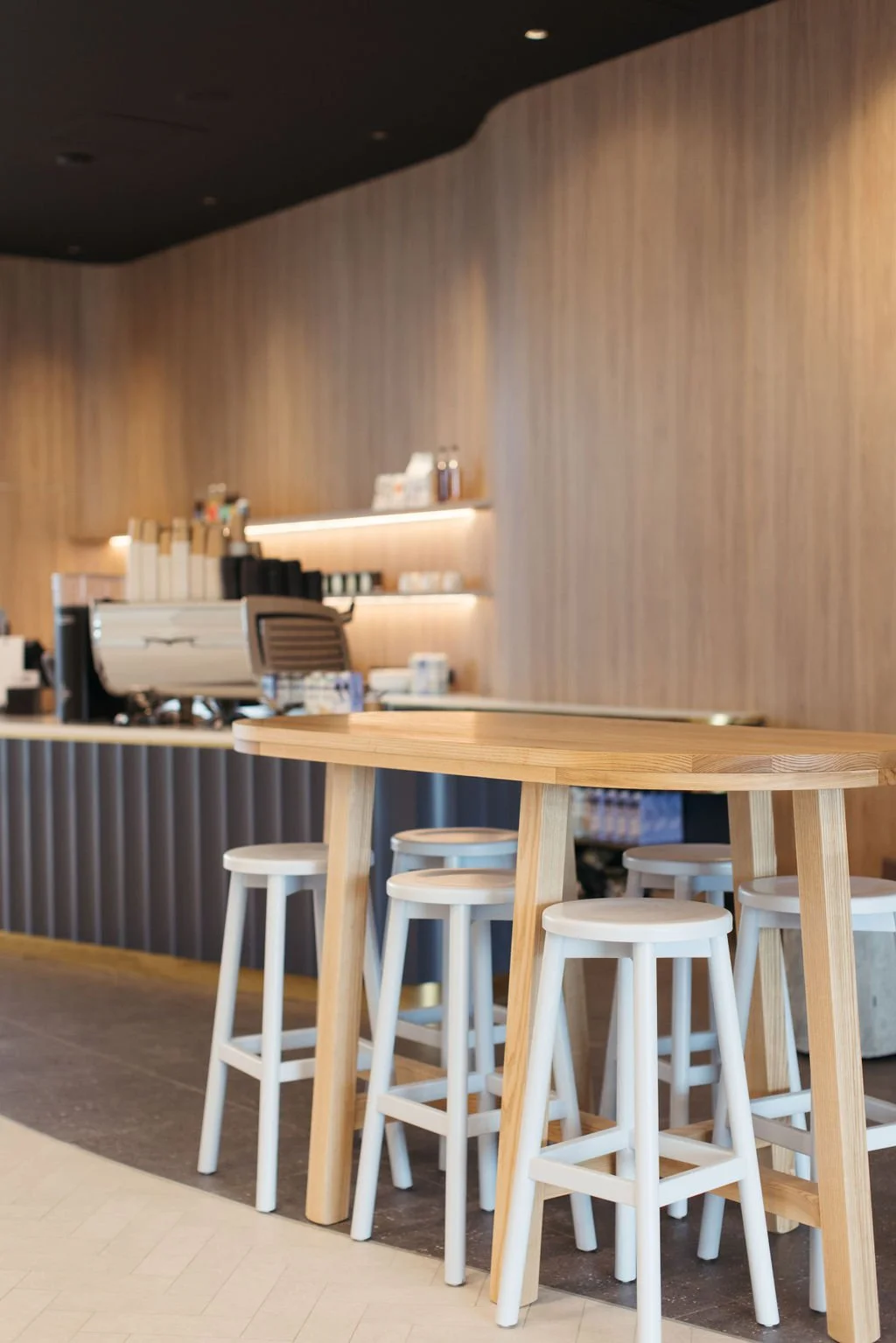 Wooden bar counter in a modern cafe featuring white stools and a background with shelves of cups and coffee machines.