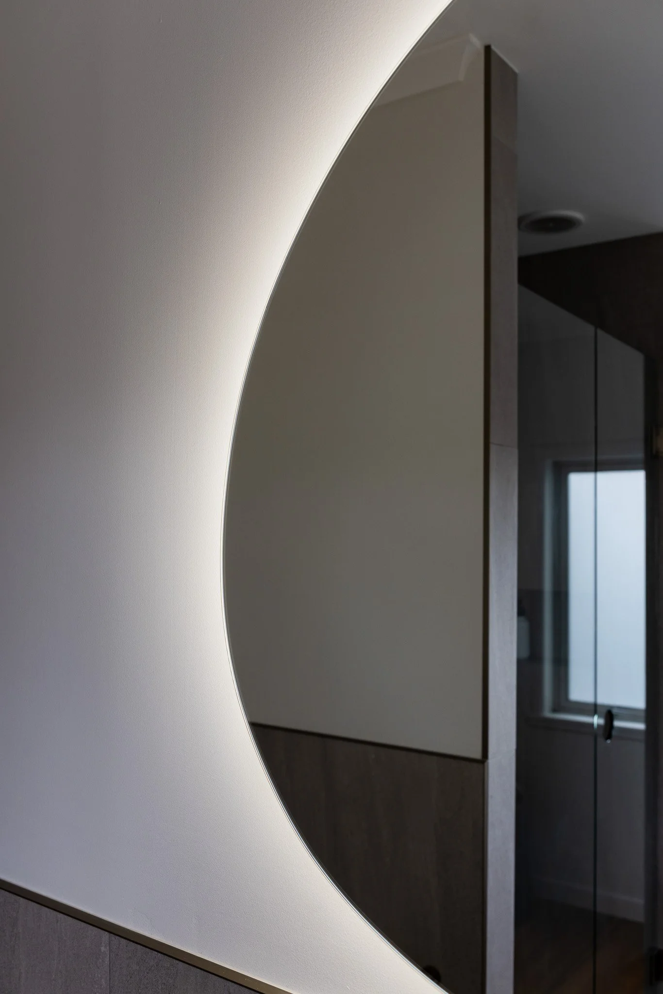 Close-up of a modern, minimalist interior featuring a curved, illuminated wall with a mirror reflecting a room with a window.
