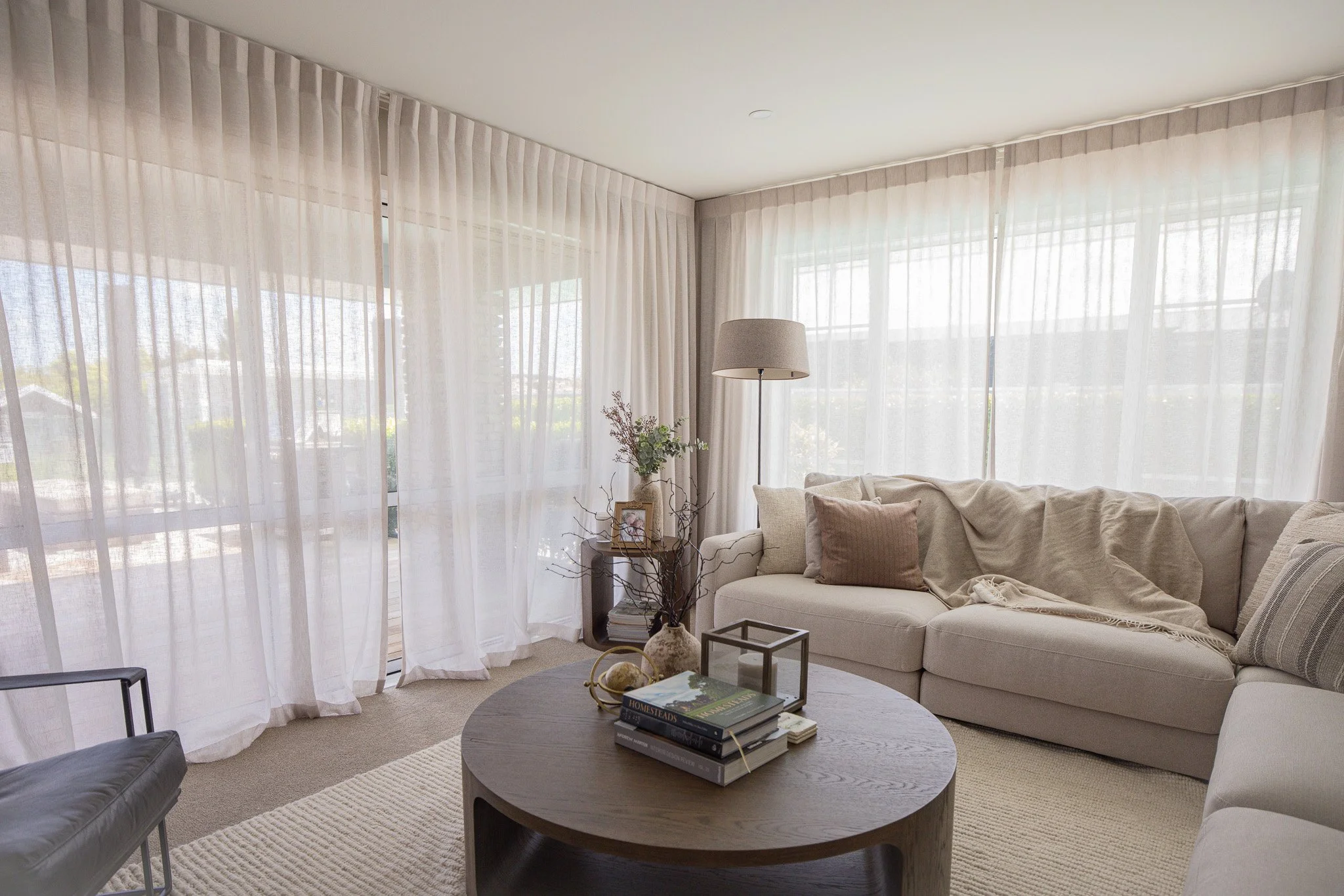Bright living room with white sectional sofa, beige pillows, and sheer curtains letting in natural light. Coffee table with books, decorative items, and a vase with flowers.