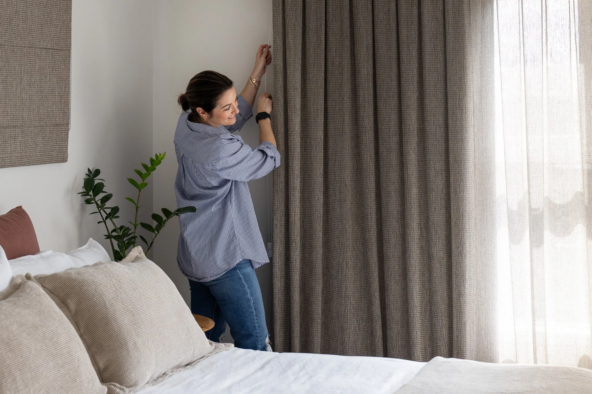 Woman adjusting curtains in a bedroom with beige pillows and a potted plant.
