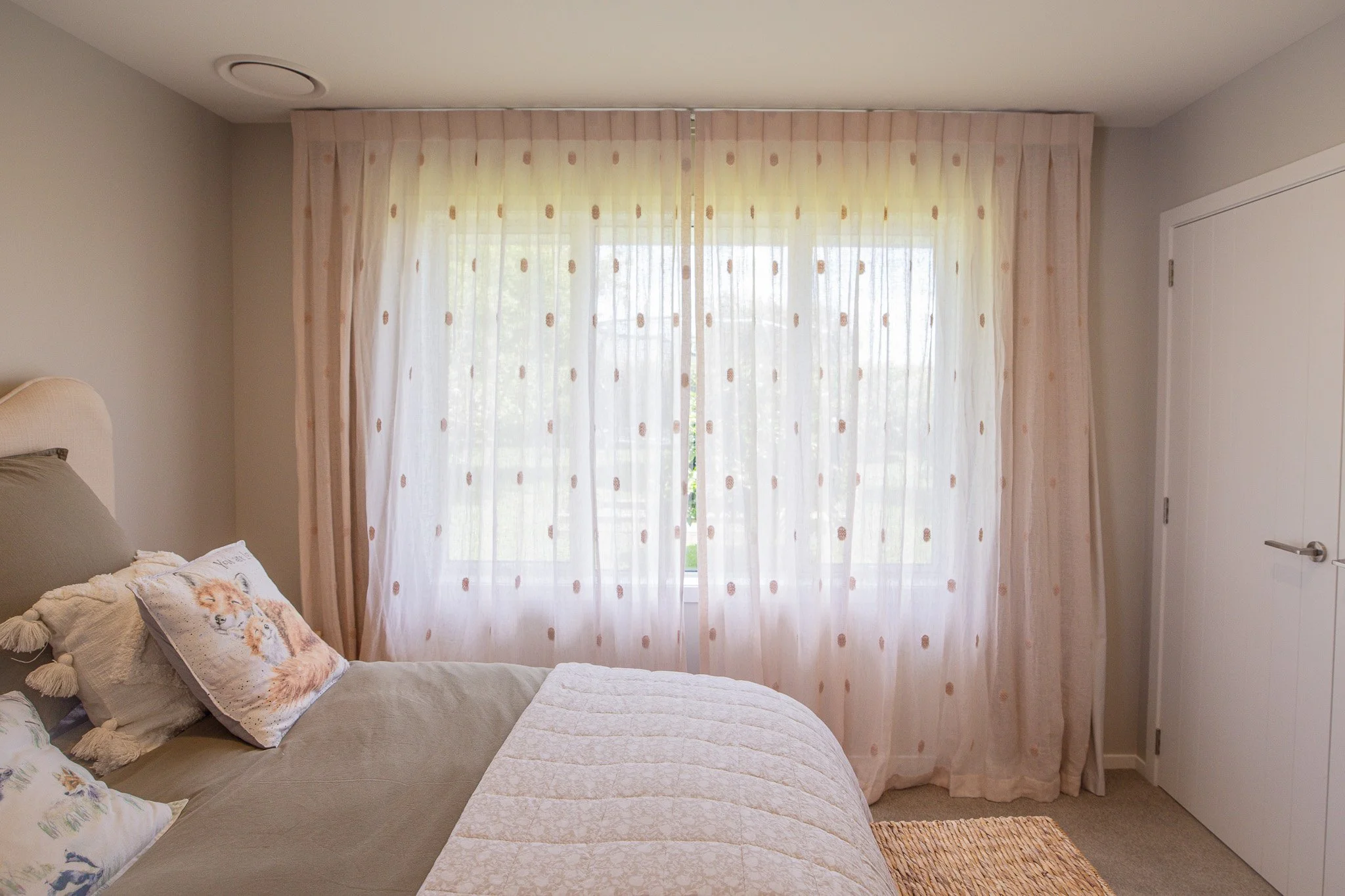 A bedroom with beige walls, a bed with gray and decorative pillows featuring animal designs, a large window with sheer pink polka-dotted curtains, and a white closet door.
